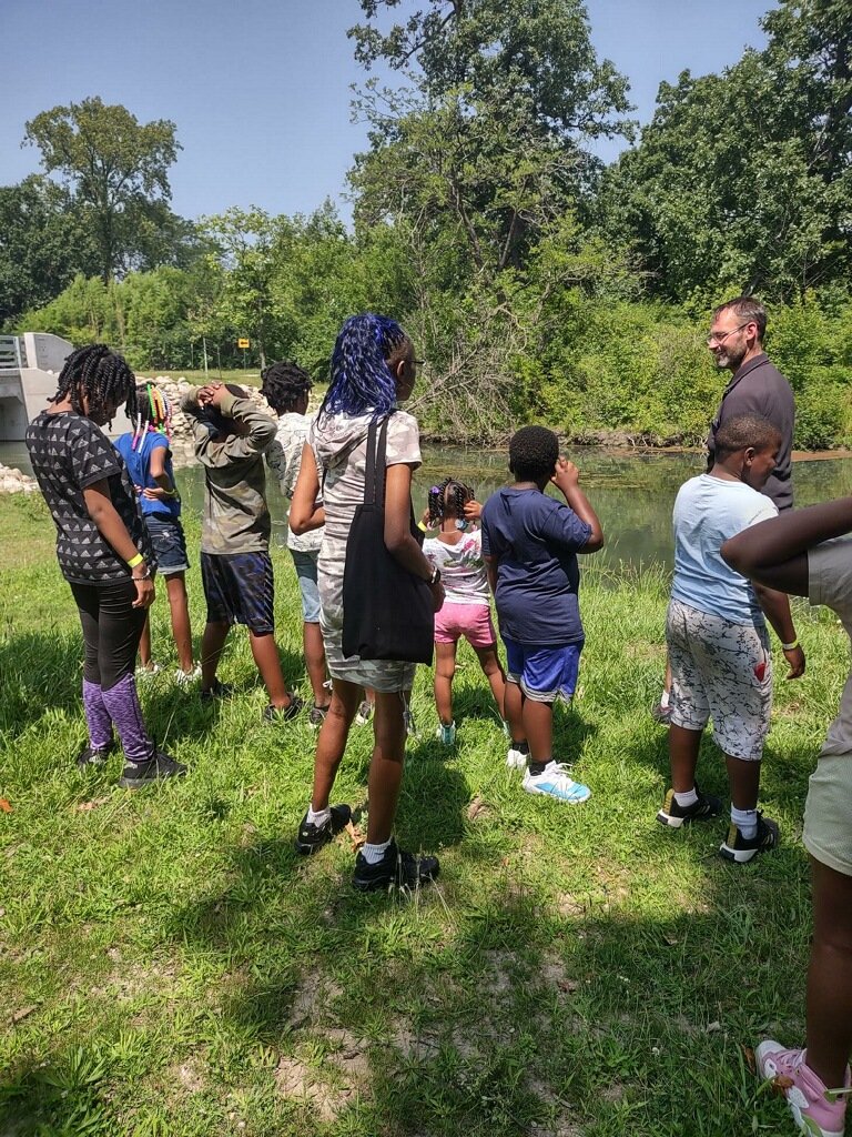 Youth participants listen to an adult beside a pond.