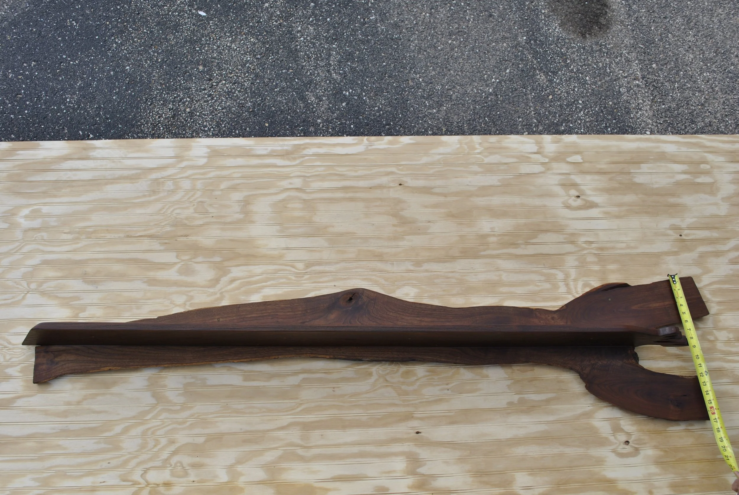 A wooden sculpture resembling a rifle with a natural, uneven shape, placed on a light wooden surface with a measuring tape next to it.