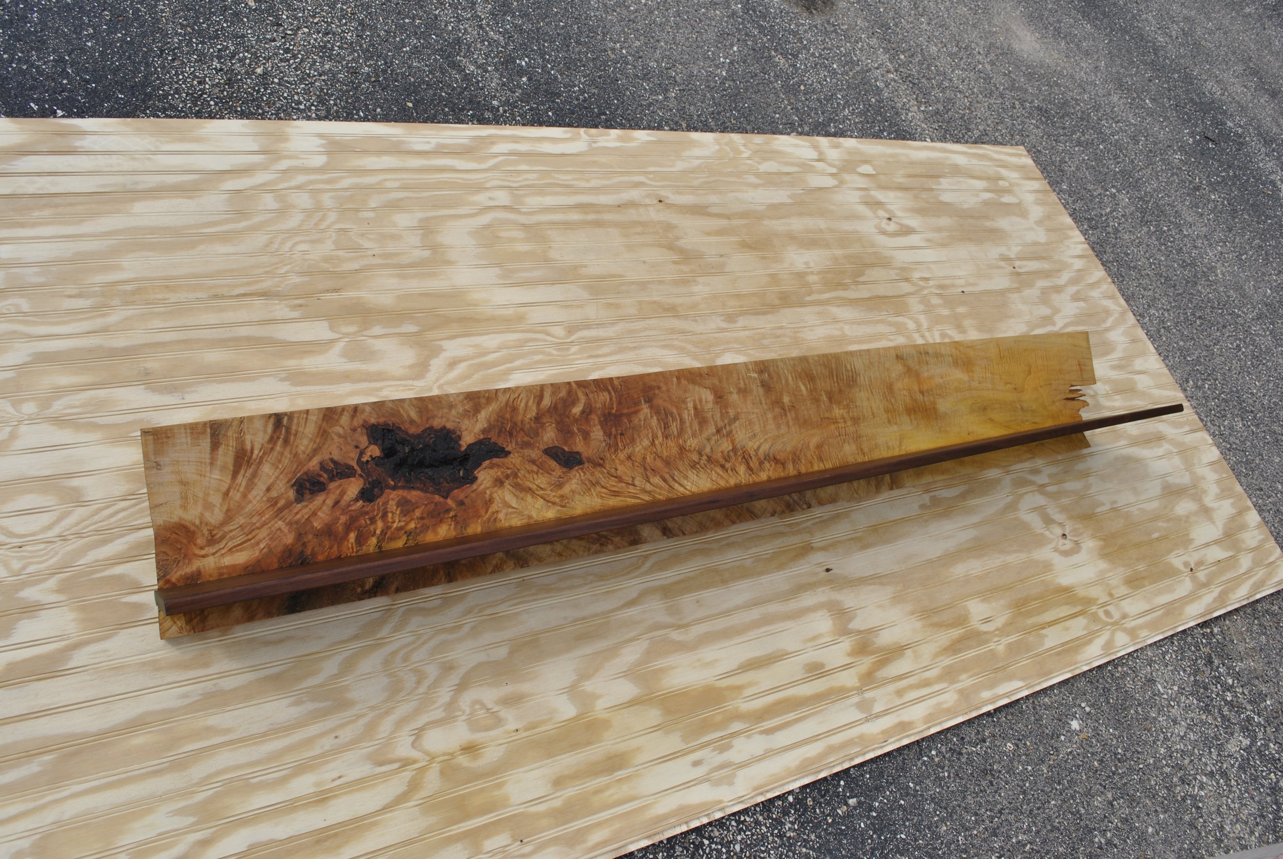 Wooden board with a section of burl wood design in the center, placed on a sheet of light-colored plywood on a gravel surface.