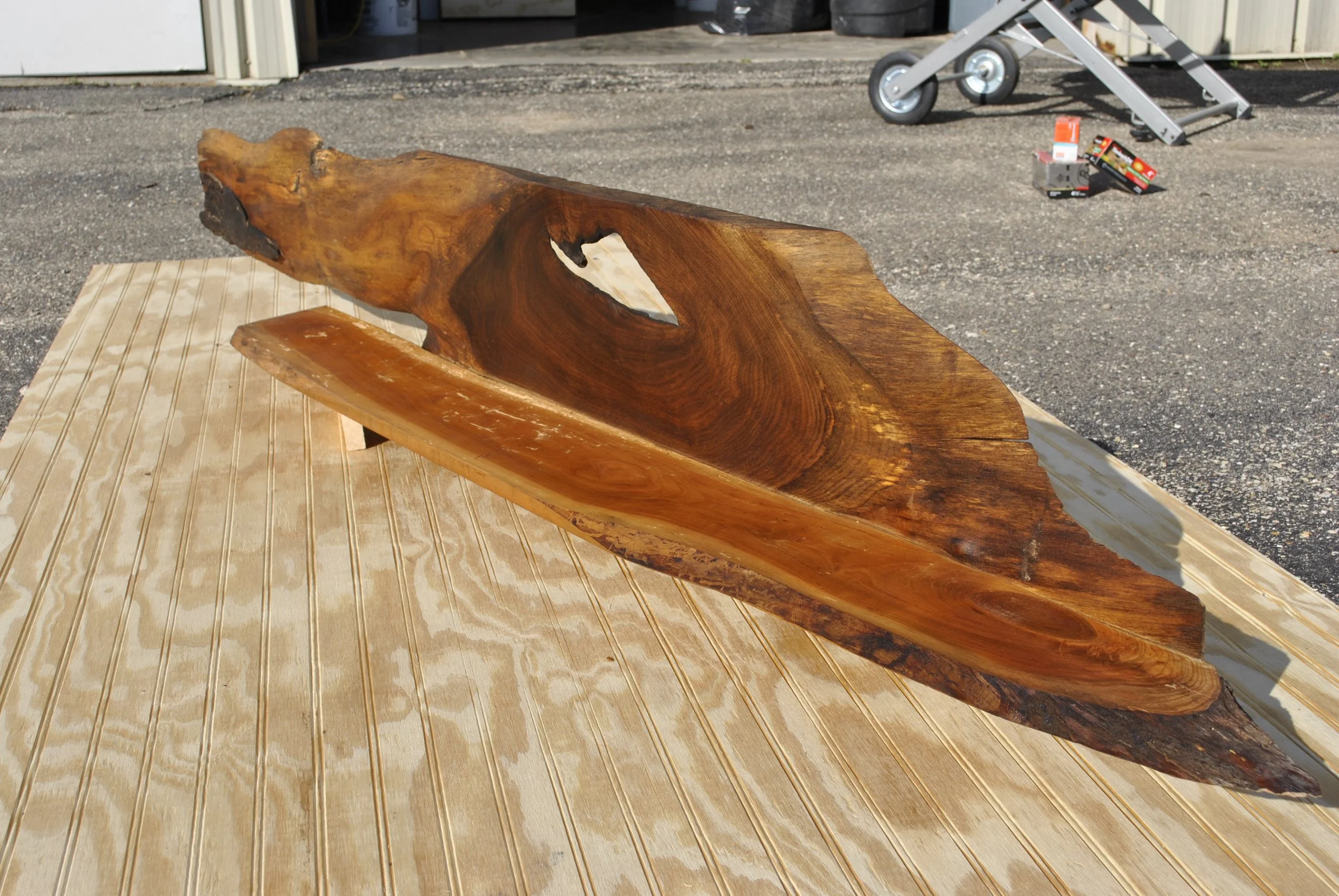 A large, polished wooden slab with a natural, irregular shape, resting on a wooden surface outside near a garage or workshop, with some tools and boxes in the background.