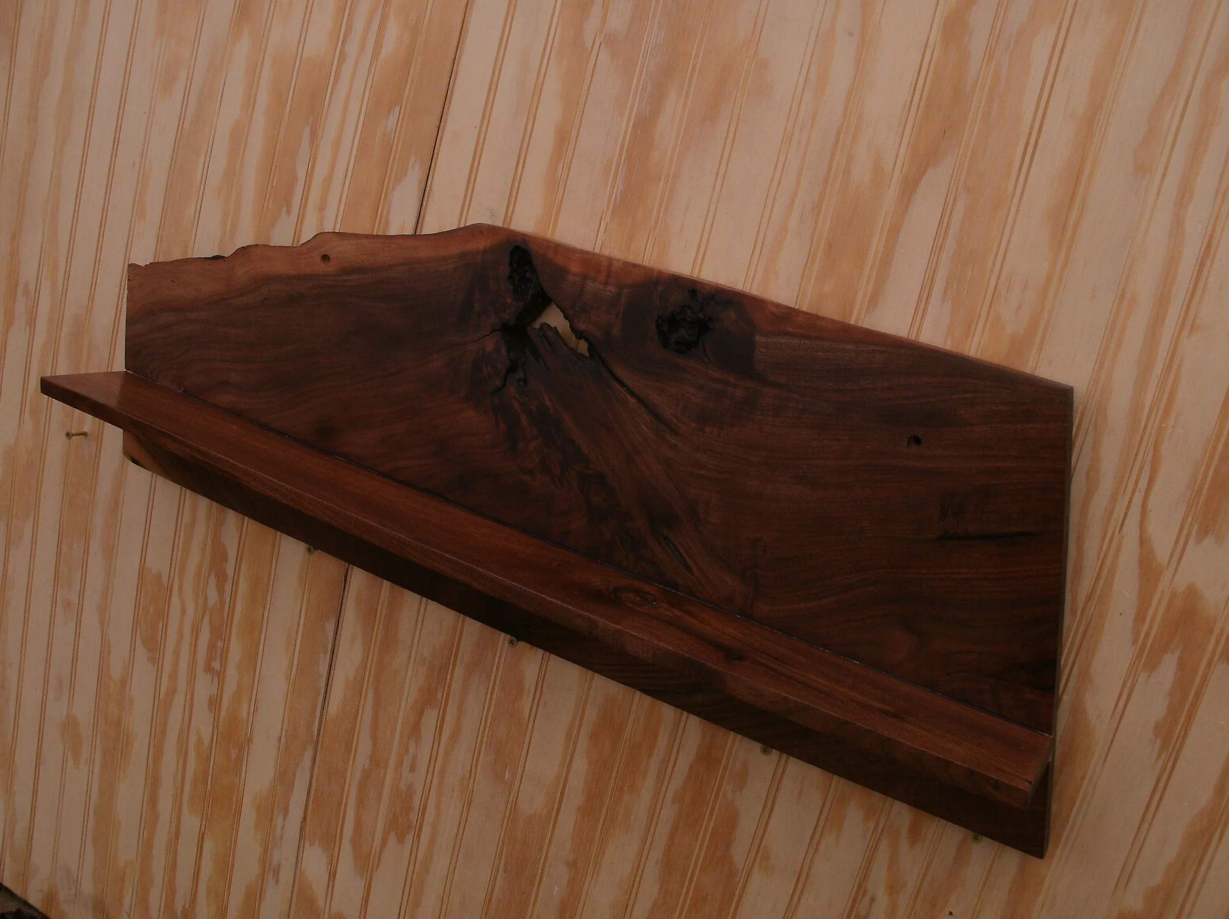 A polished wooden shelf or piece of furniture with natural wood grain and knots, mounted on a wooden wall.