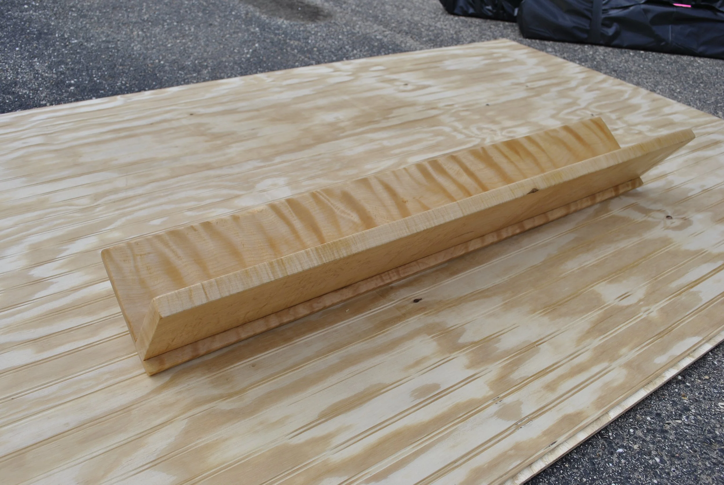 A piece of light-colored wood with a raised angled edge, placed on a large wooden surface outdoors on a rough asphalt ground.