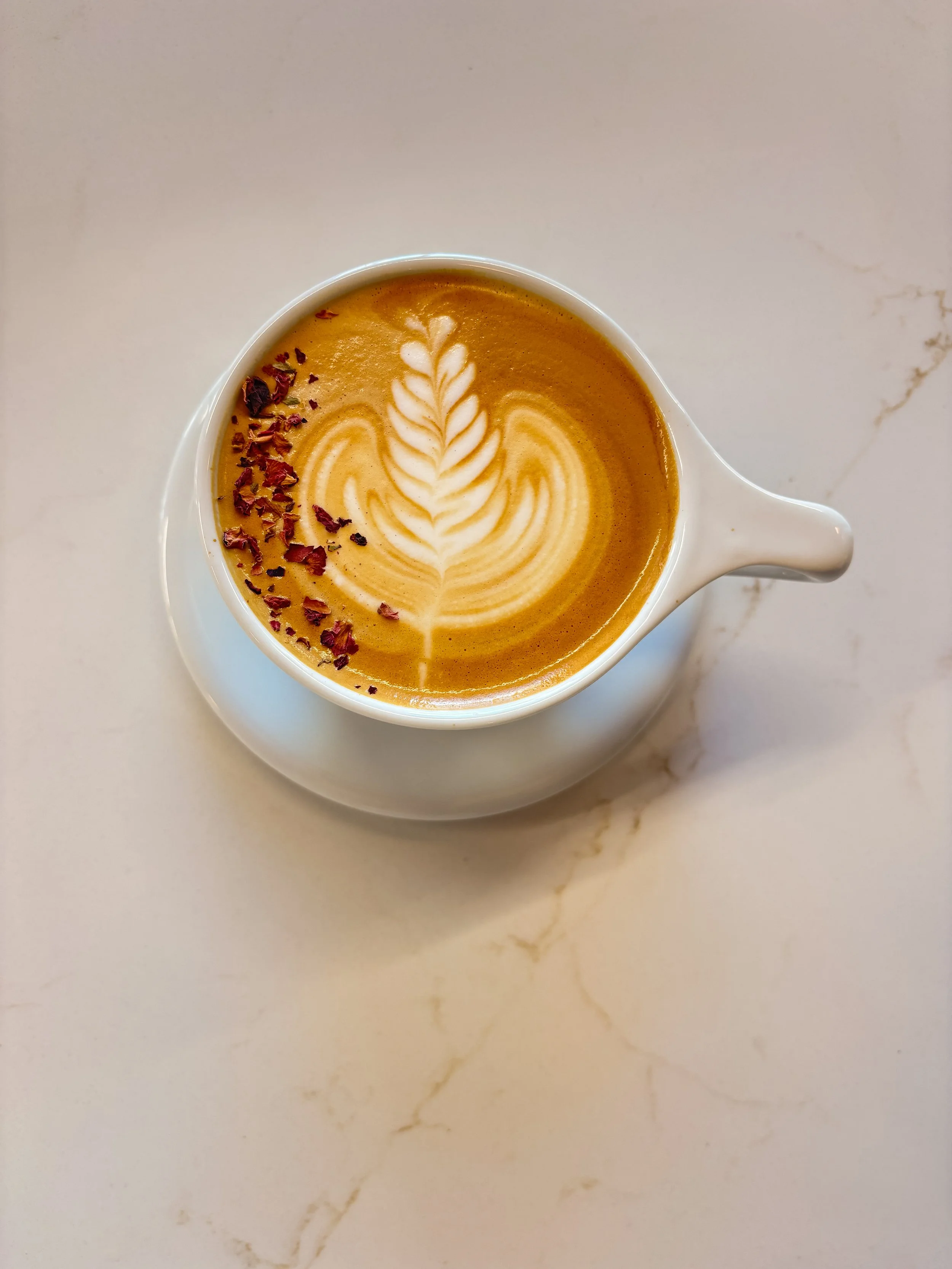Signature rose latte in a round white mug, with rosetta latte art and sprinkled with rose petals on marble countertop.