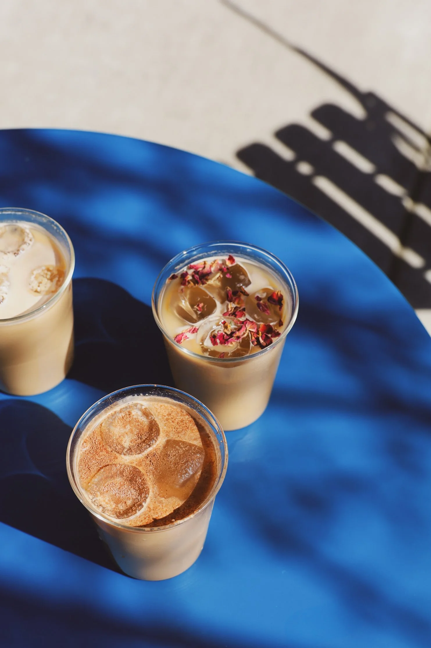 Three iced coffee drinks in plastic cups on a blue table, one garnished with rose petals.
