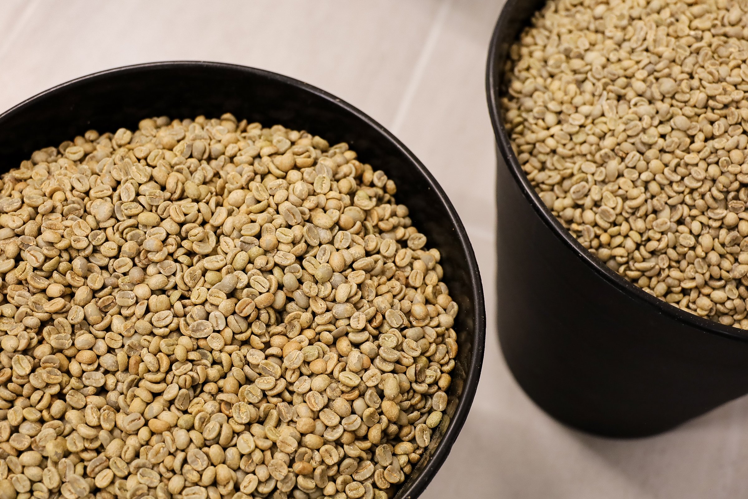 Two black bowls filled with raw green coffee beans, positioned on a light surface.