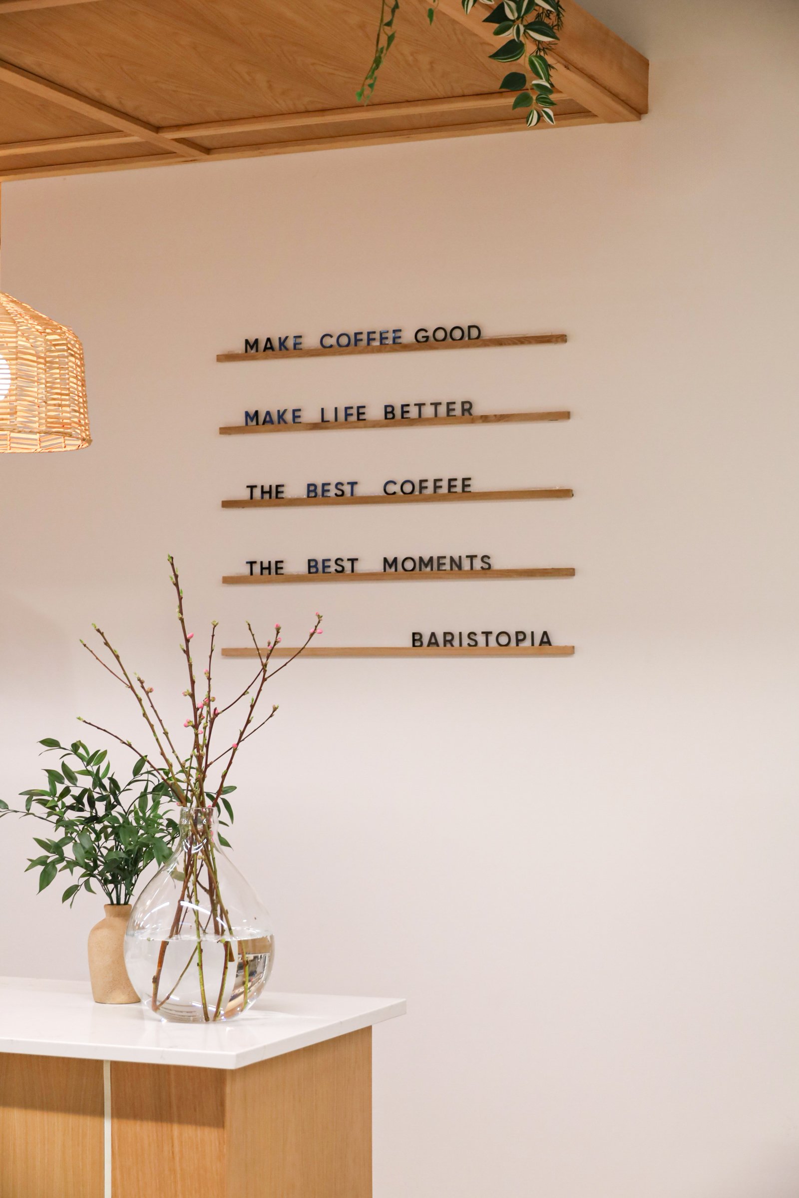 Decorative coffee shop interior with motivational wall quotes, a wooden ceiling, wicker light fixture, and a glass vase with green plants and branches on a countertop.