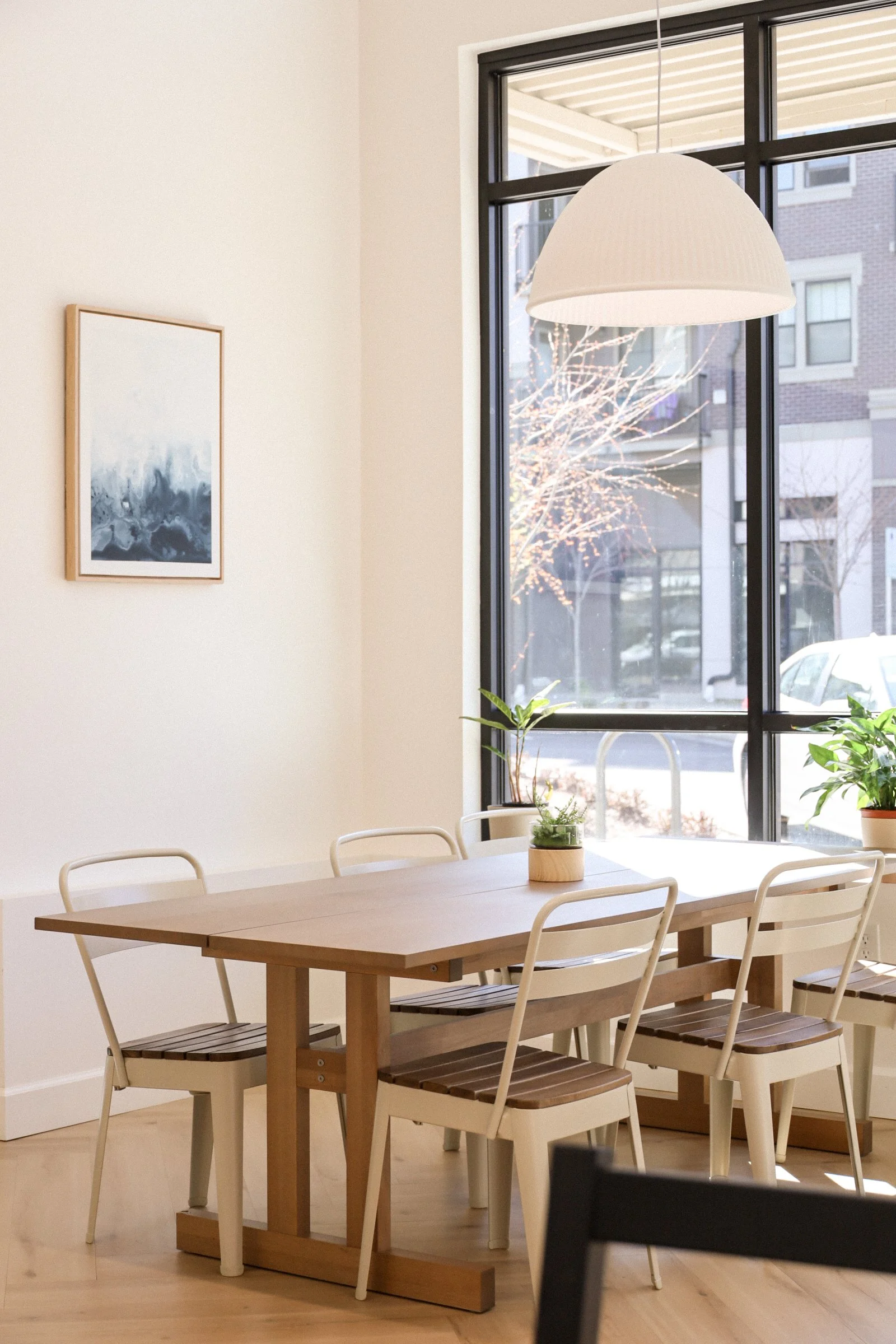 Bright minimalist dining area with wooden table, white chairs, indoor plants, large window, modern pendant light, and wall art.