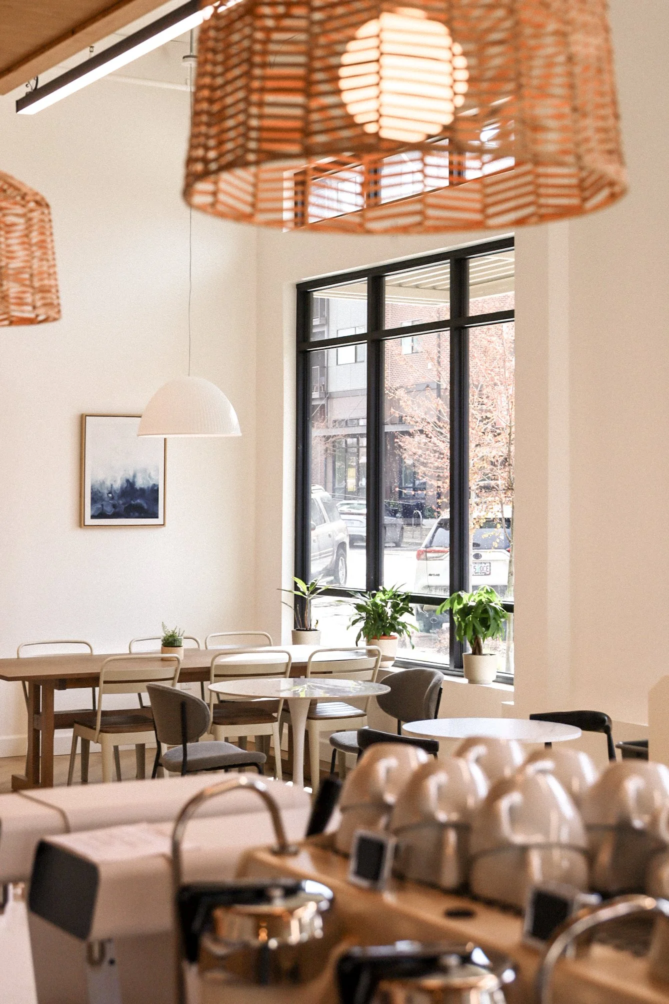 Bright and modern cafe interior with large windows, wooden tables, chairs, decorative plants, pendant lights, and coffee equipment in foreground.