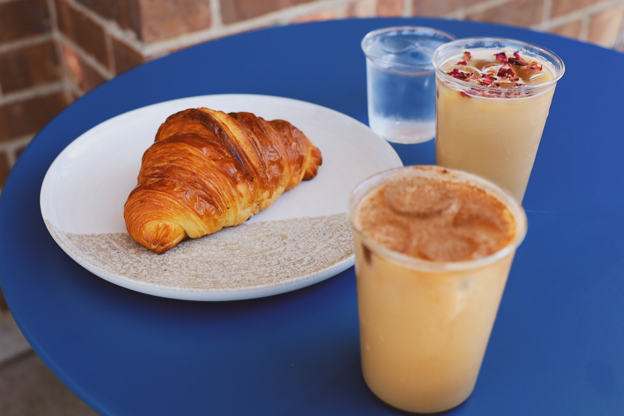 Croissant on a plate with two iced drinks, one with foam and petals, on a blue table.