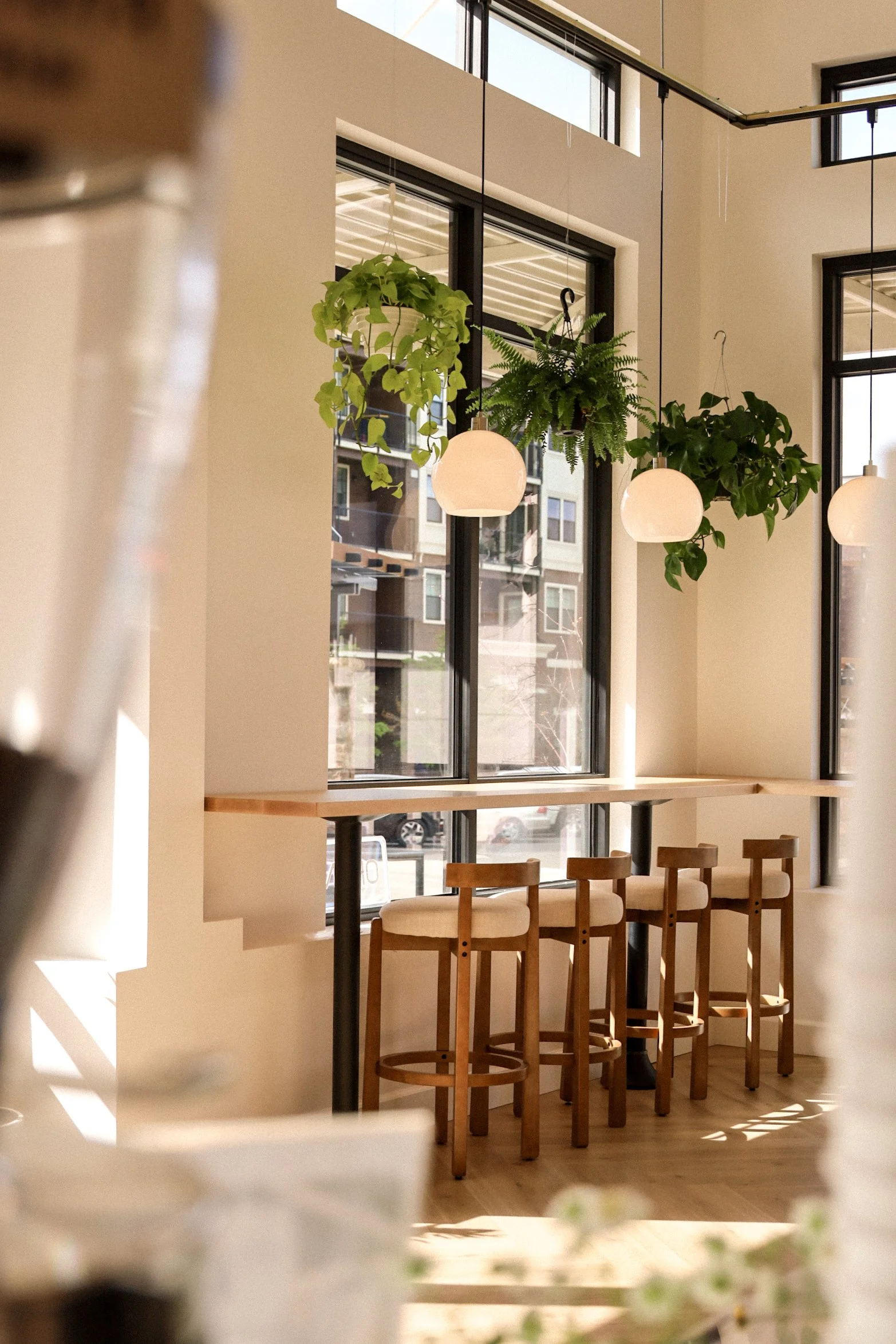 Interior of a modern cafe with wooden stools and high bar table near large windows, hanging plants and pendant lights