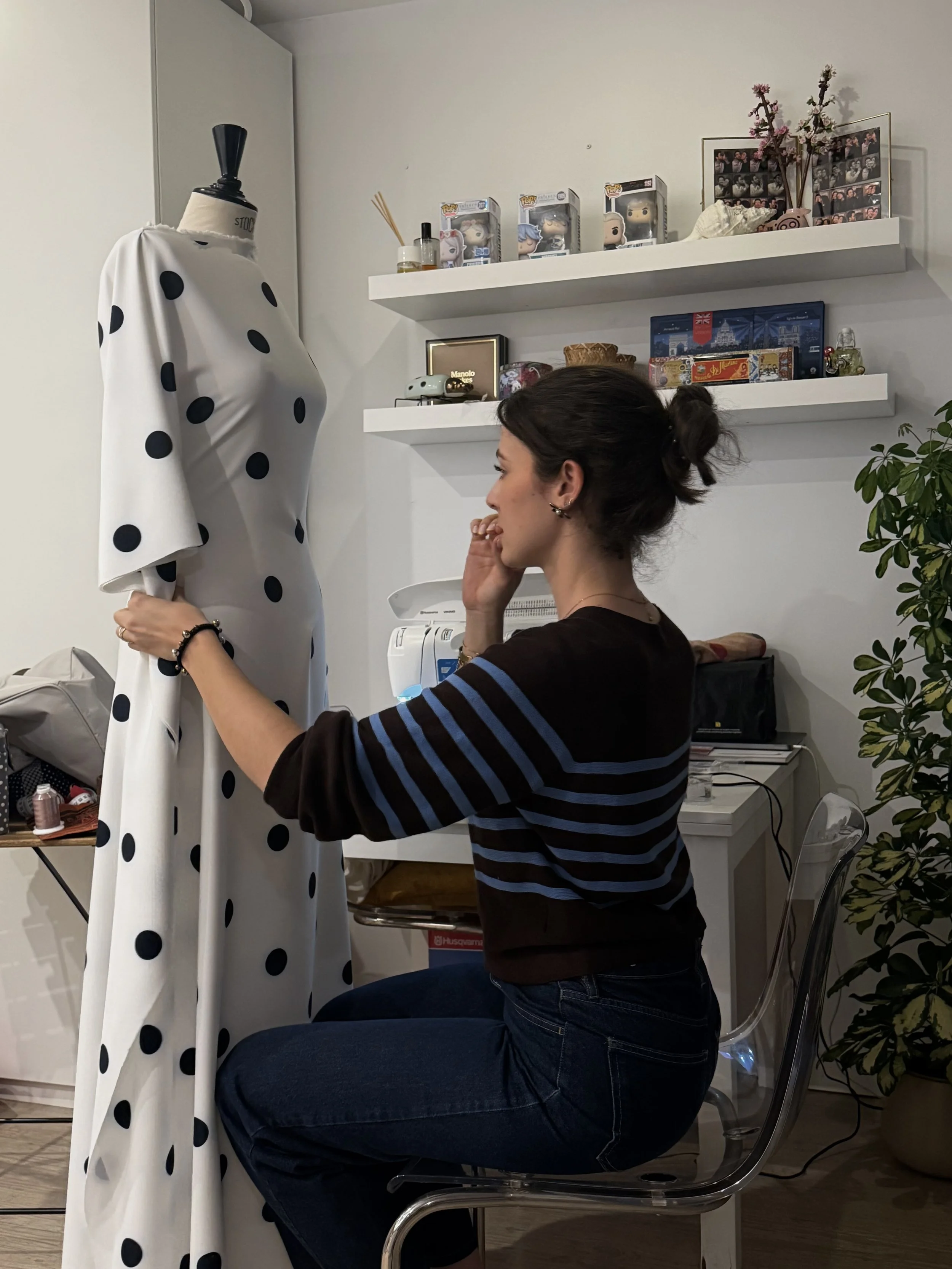 Mujer sentada en una silla transparente, viendo a una figura de tela con puntos negros, doblando esa figura, en una habitación con estanterías con juguetes, libros y decoraciones como fotos y plantas.