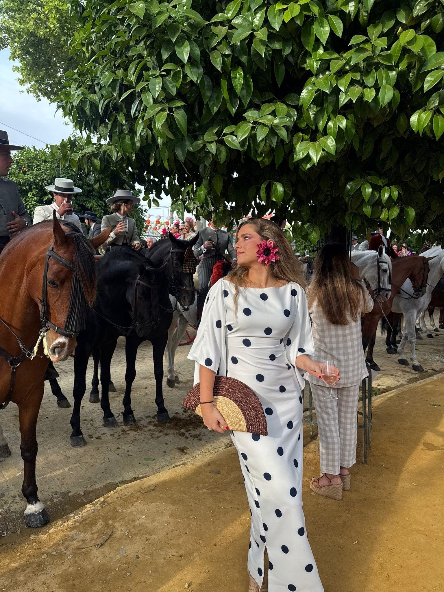 Una mujer con vestido blanco de puntos negros, con una flor rosa en el cabello, sosteniendo un sombrero y una copa de vino, en un evento con caballos y personas vestidas elegantes.