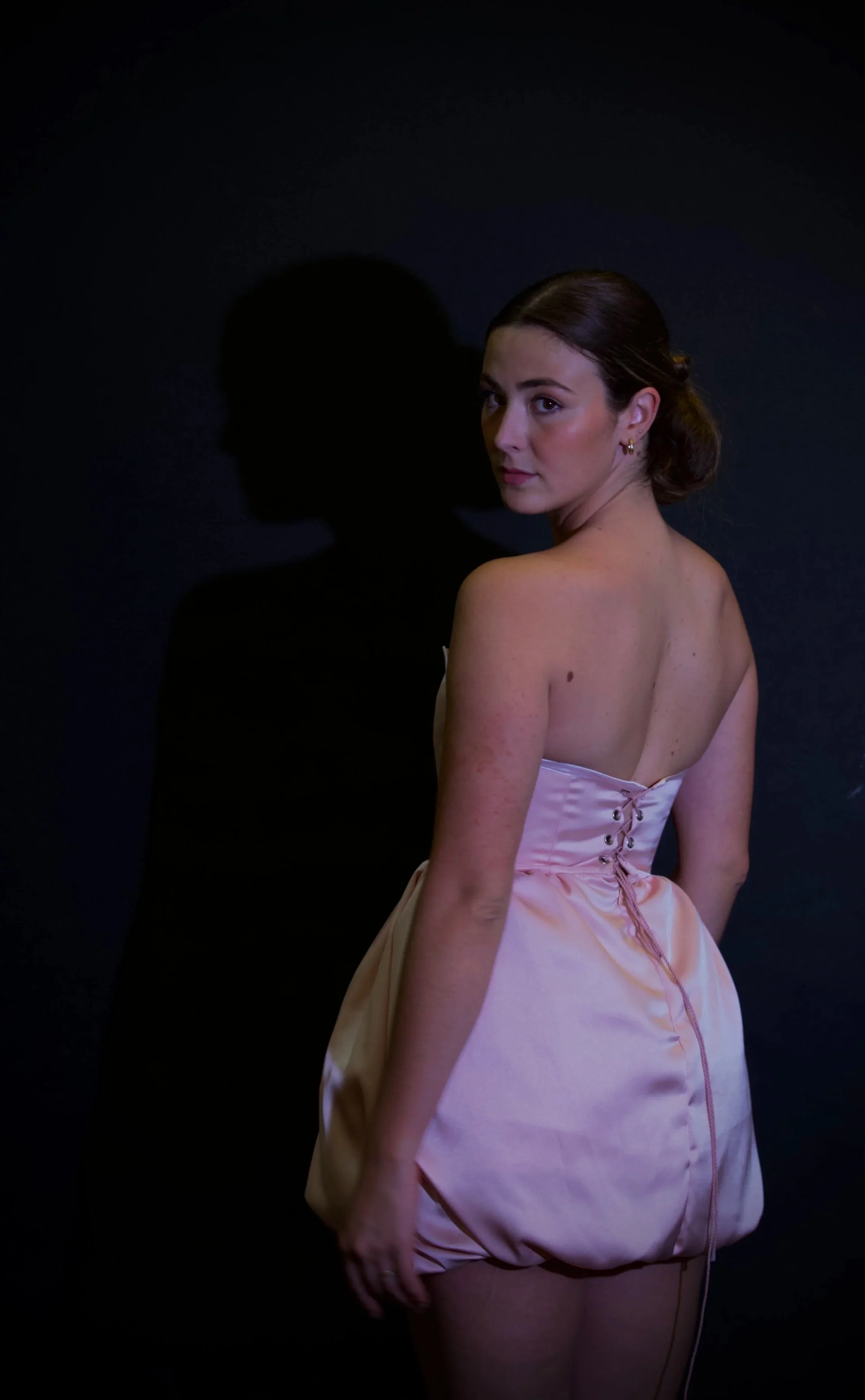 Mujer con vestido rosa y cabello recogido posa frente a un fondo oscuro, con una sombra proyectada en la pared a su izquierda.