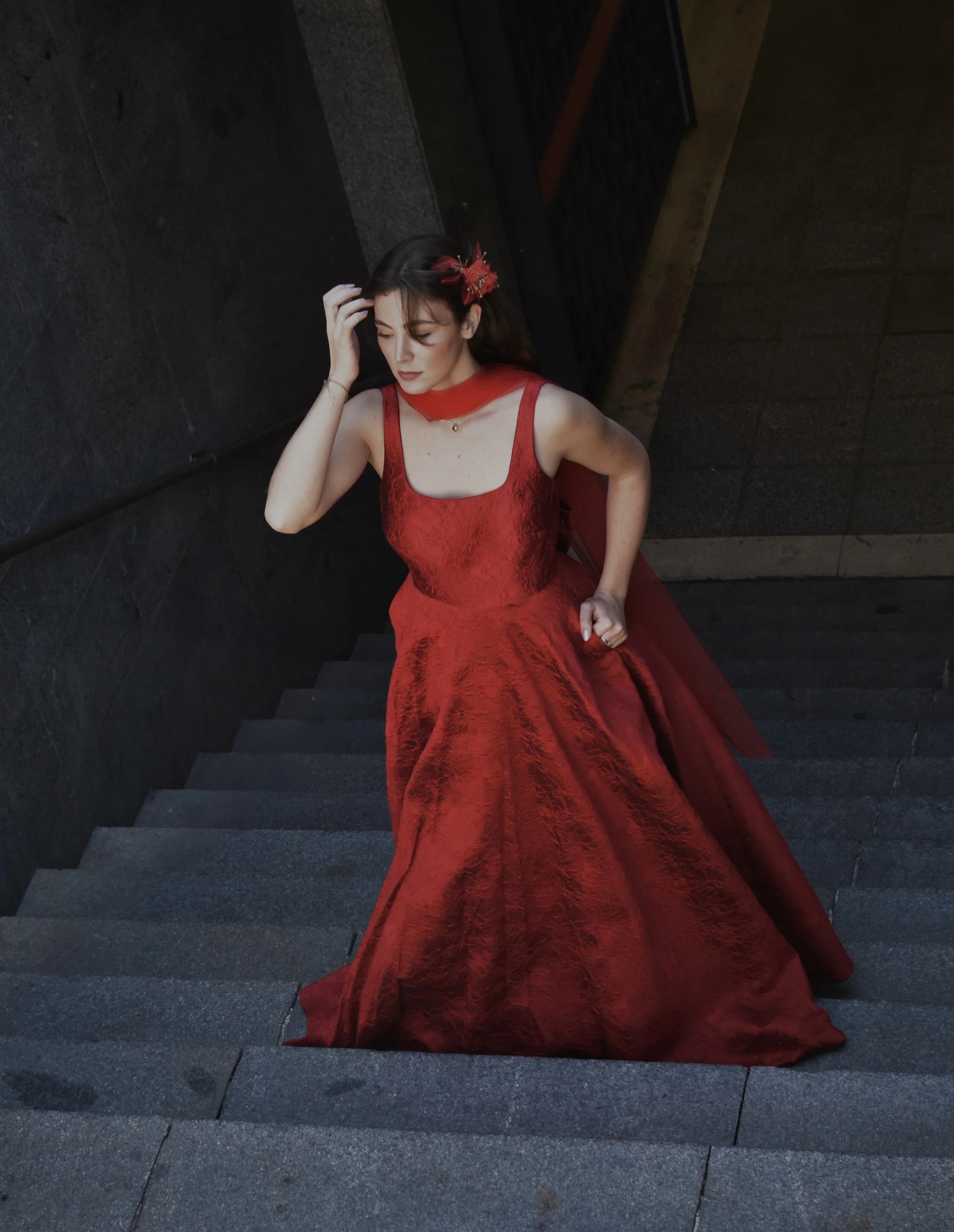 Mujer con vestido largo y rojo en una escalera, parece pensativa.