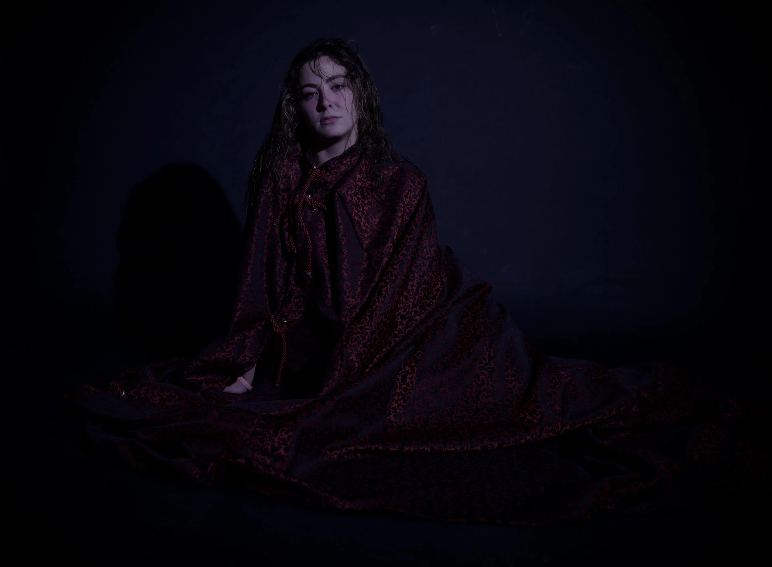 Mujer con vestido largo de color oscuro, sentada en el suelo, con cabello rizado y expresión seria, en un fondo negro.