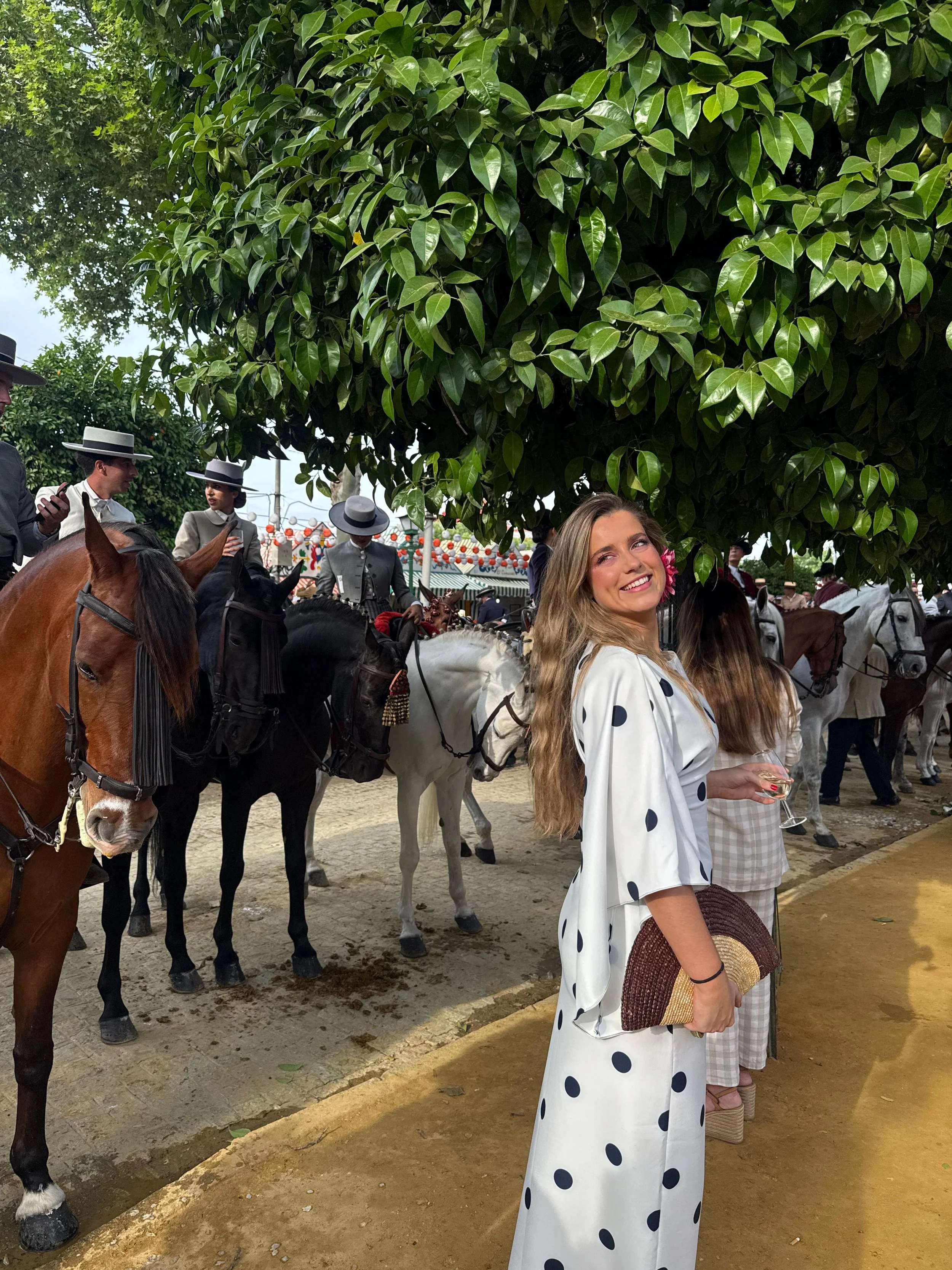 Mujer sonriendo con vestido blanco con puntos oscuros, sosteniendo un sombrero en una mano y una copa en la otra, rodeada de personas a caballo en un evento al aire libre con árboles y decoraciones de linternas.