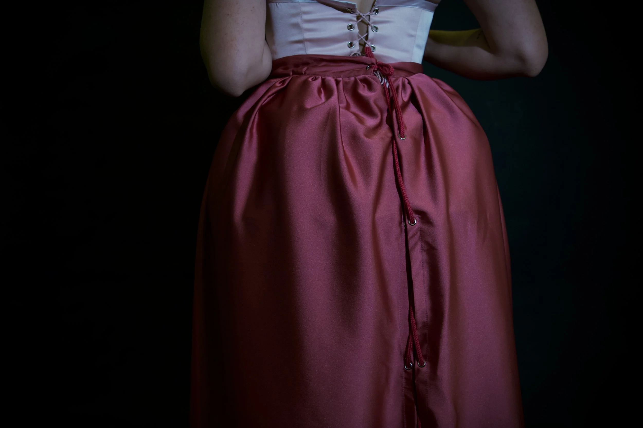 Parte superior de una persona con un vestido de corsé blanco y falda larga de satén rojo, contra fondo oscuro.
