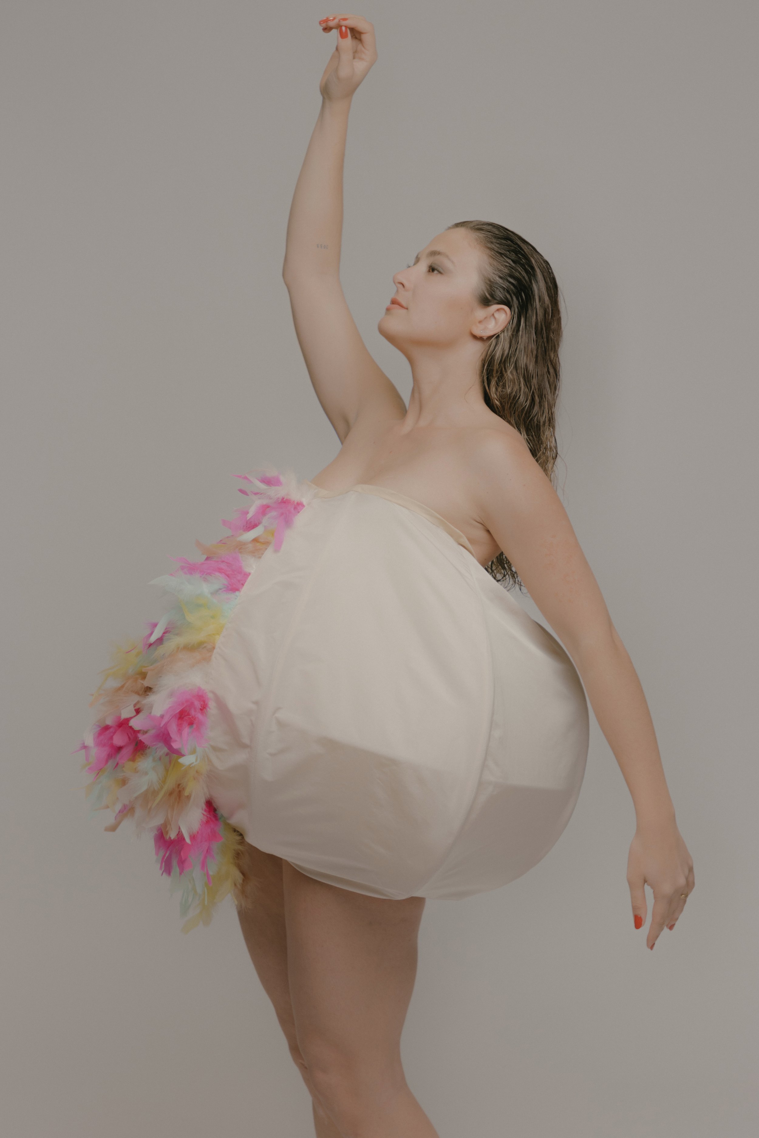 Mujer desnuda con un vestido de burbuja y decoración de plumas, posando con los ojos cerrados y brazo levantado.