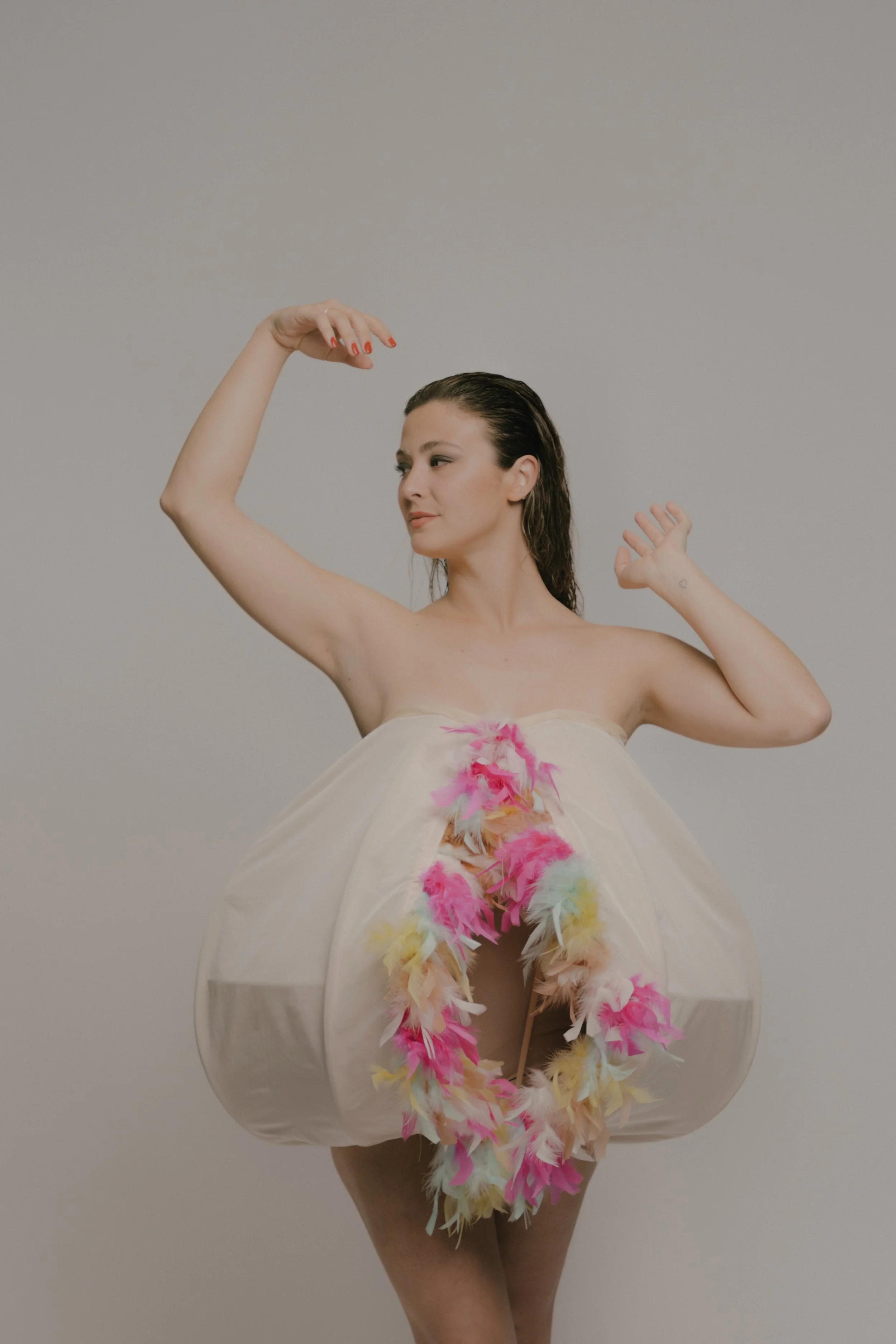 Mujer desnuda con maquillaje natural y cabello húmedo, adornada con plumas de colores pastel en un vestido transparente y falda, posando frente a un fondo gris.