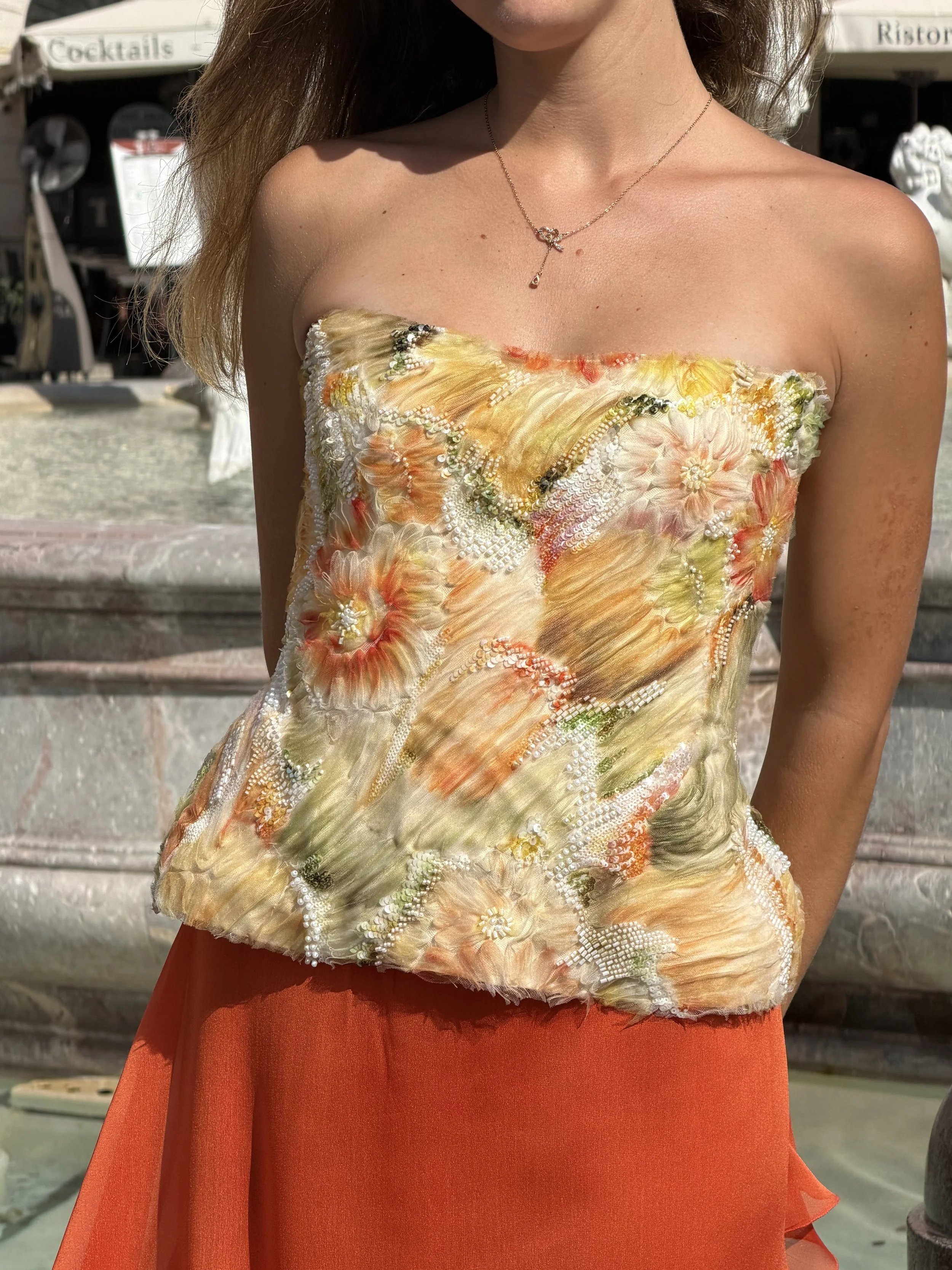 Mujer con blusa strapless con detalles de bordado y perlas, luciendo un collar dorado delgado con un pequeño colgante, en un ambiente soleado.