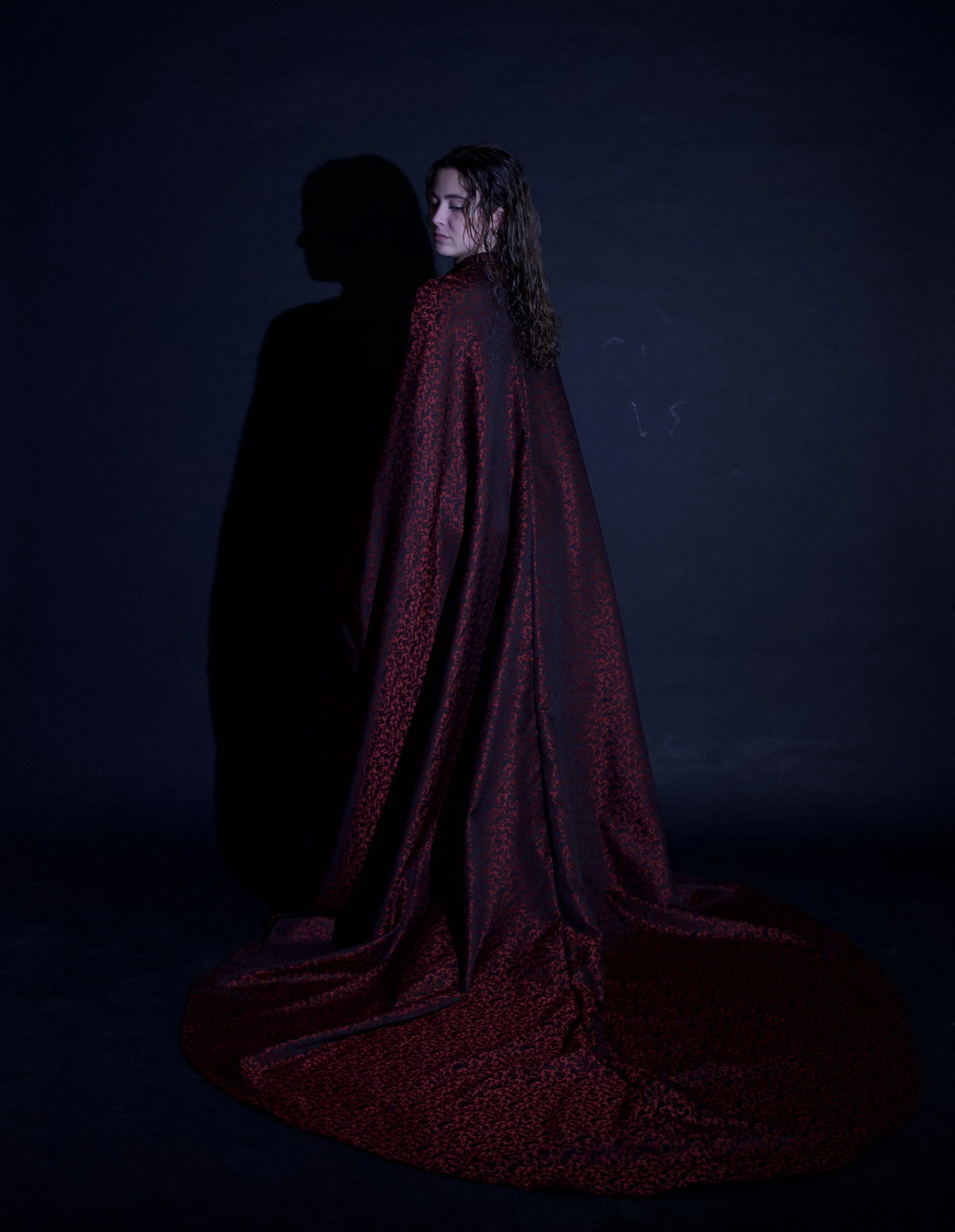 Mujer con cabello rizado y piel clara, vestida con una capa larga y elegante, de color rojo oscuro con patrones, de pie contra una pared azul oscuro, proyectando su sombra en la pared.