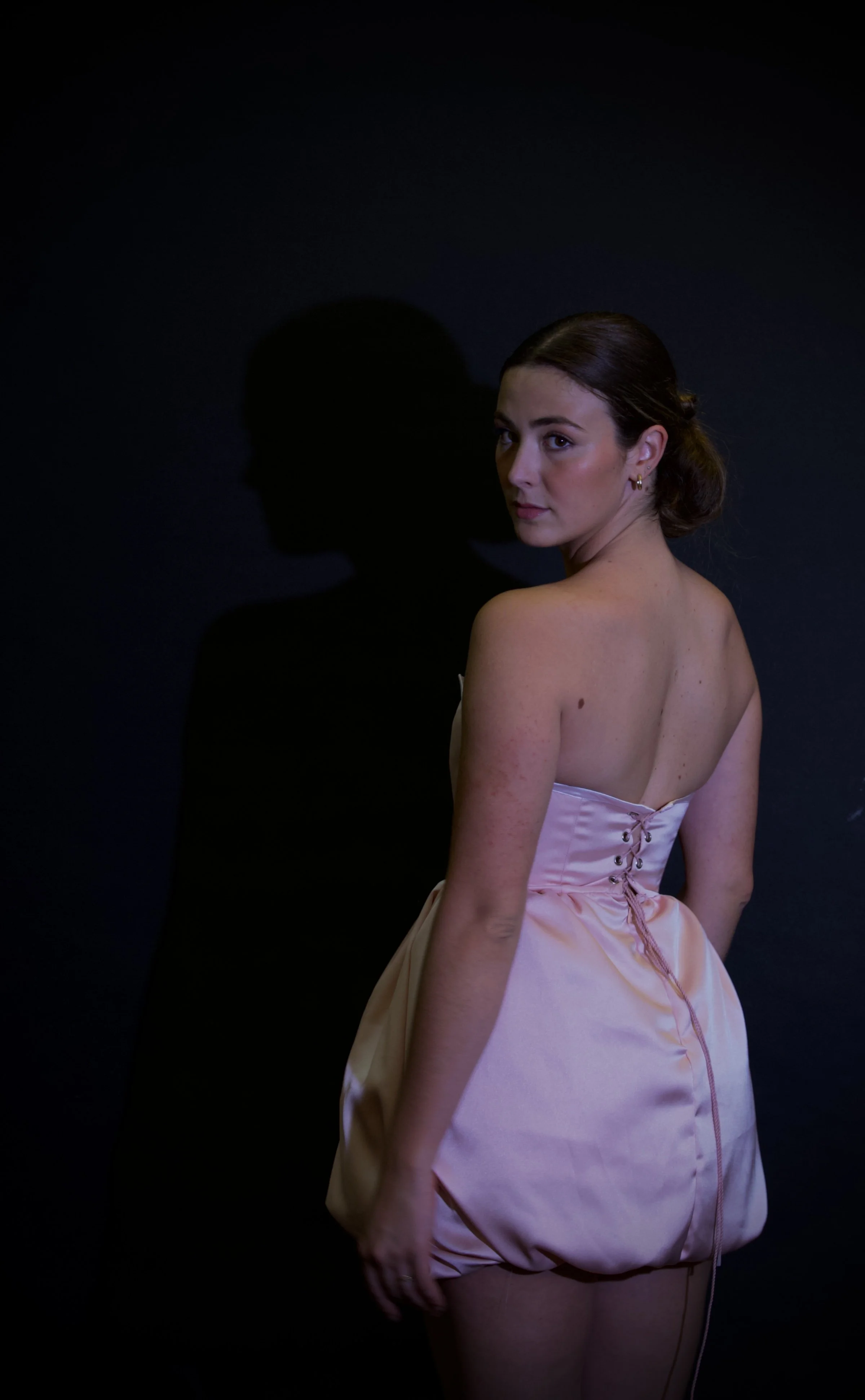 Retrato de una mujer con vestido de satén rosa, de espaldas, mirando hacia la cámara, con fondo negro y sombra de su perfil en la pared.