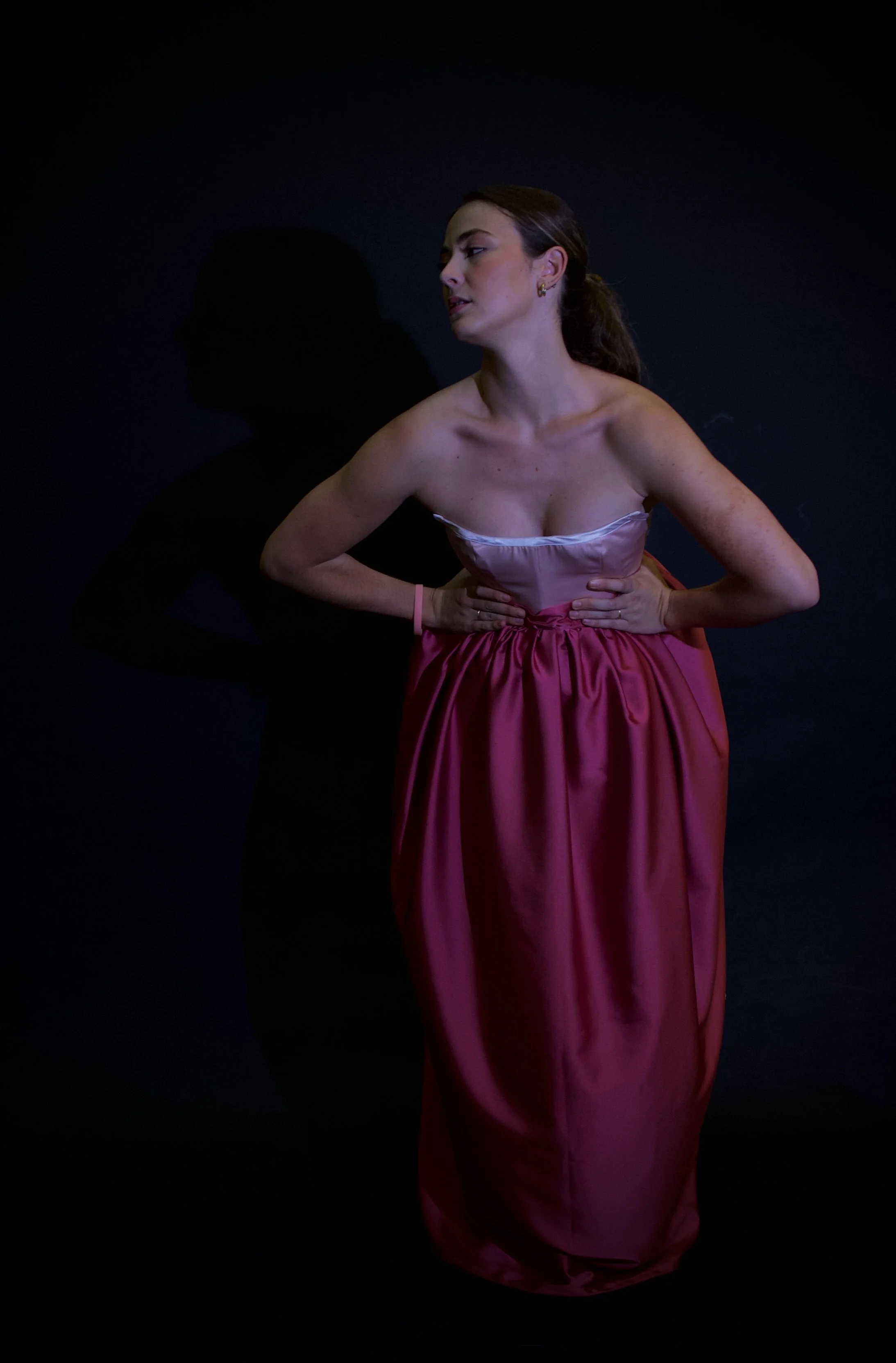 Mujer con vestido rosa y sin hombros se apoya en las caderas mientras mira hacia un lado, con sombra en la pared detrás.