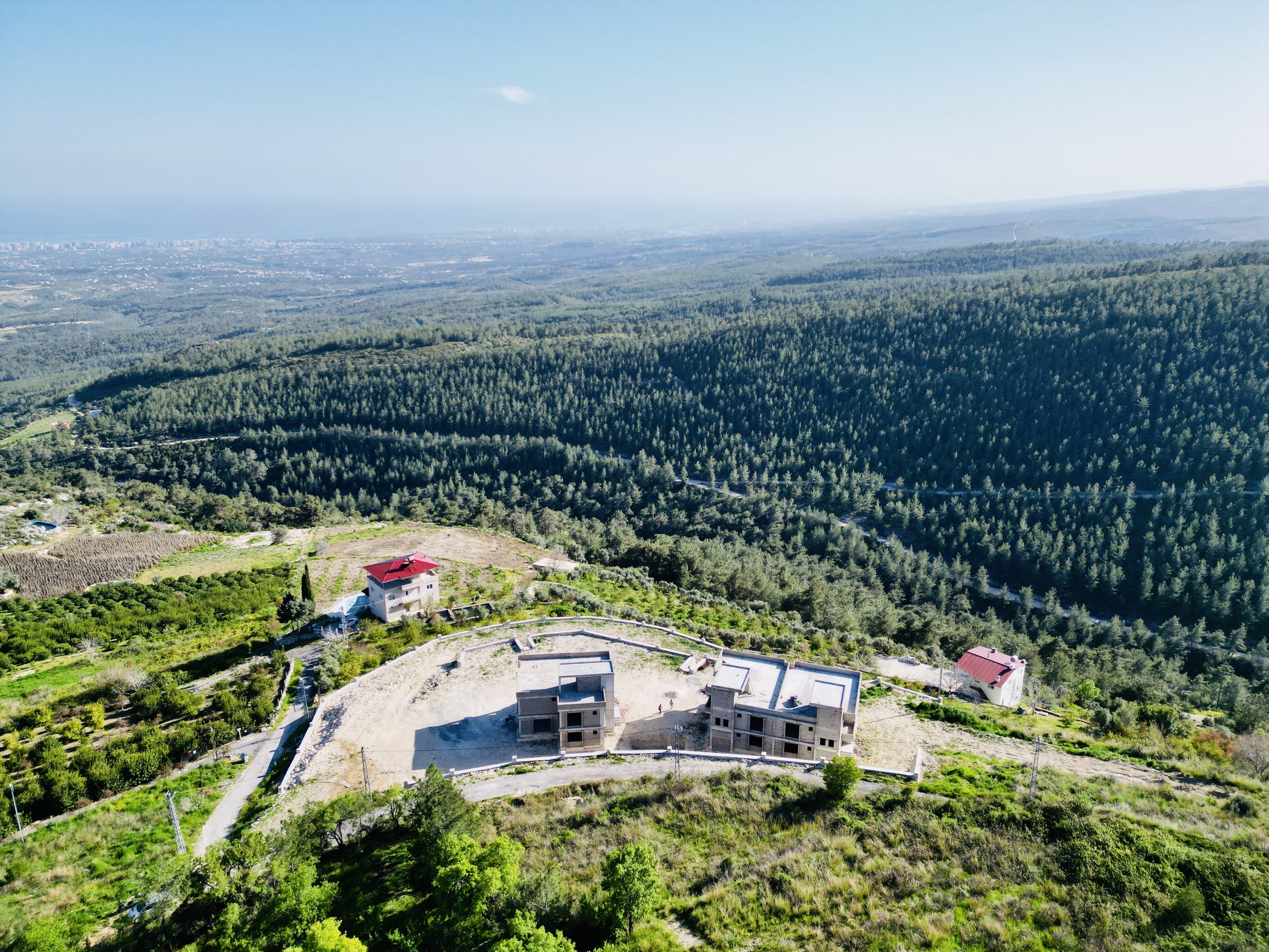 Aerial view of a wooded landscape with scattered buildings, including houses with red roofs, surrounded by dense green forests and open areas, under a clear blue sky.