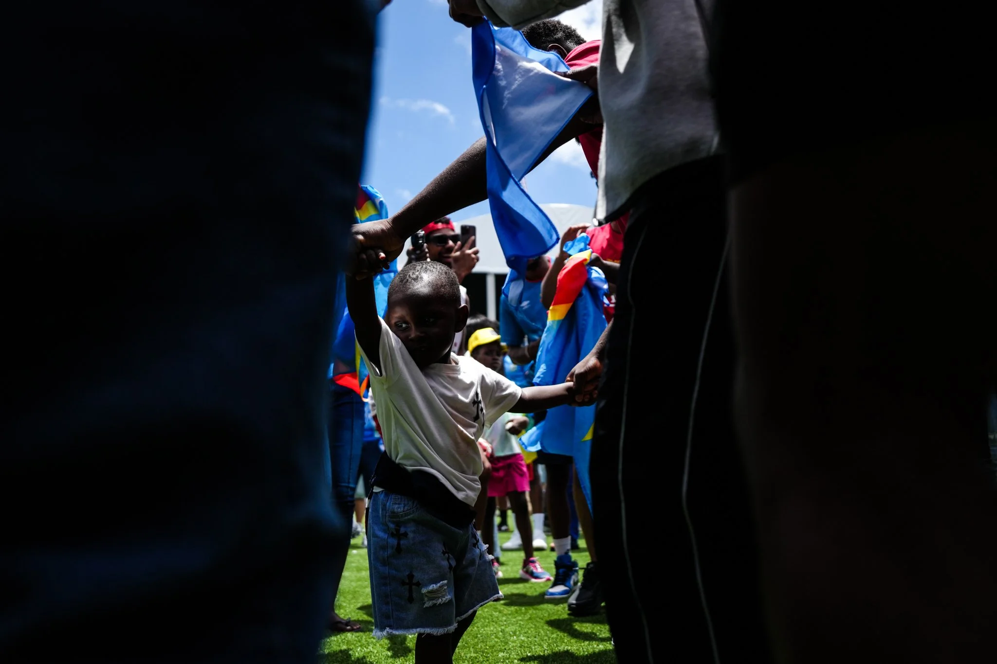 A young boy with a shaved head and dressed in a white t-shirt and jeans with patches is holding hands with an adult in a gray jacket, standing on a grassy field during daytime. Several people are standing in a line behind them, some wearing colorful 