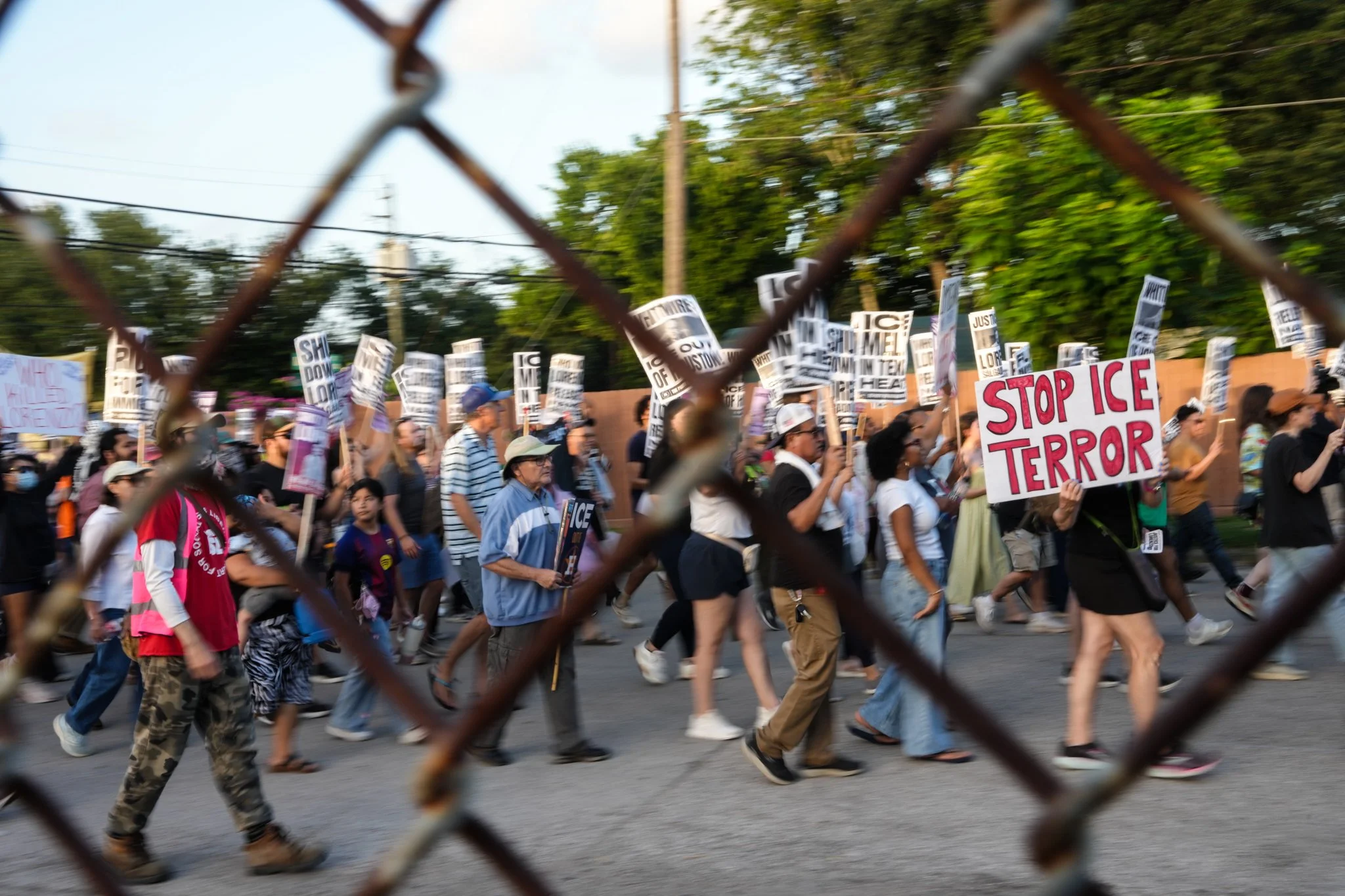 Protestors holding signs with messages such as 'Stop ICE Terror' and other signs, marching on a street seen through a chain-link fence, with trees and utility poles in the background.