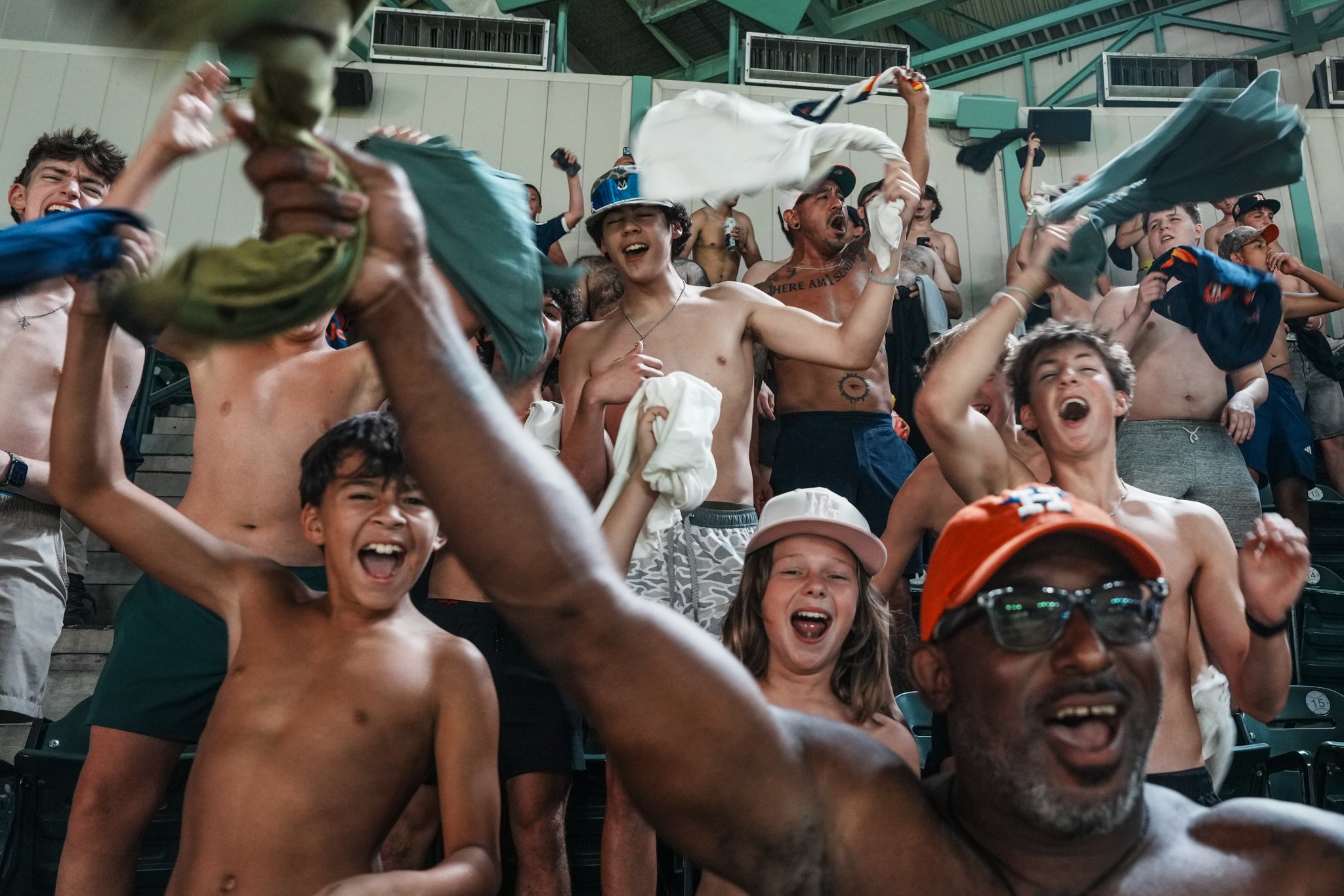 A group of young people cheering and celebrating, some shirtless, at an indoor event with wooden tiers and green beams.