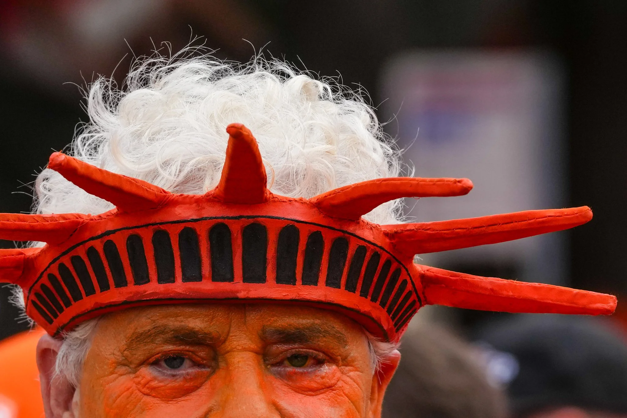 Close-up of a person dressed as the Statue of Liberty, wearing a red headpiece with spikes and a curly gray wig, with visible wrinkles and gray eyes.