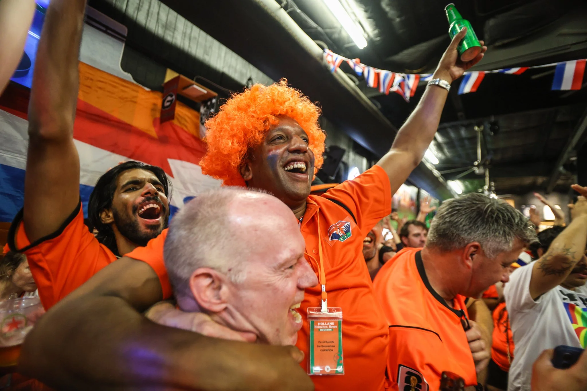 Crowd of enthusiastic sports fans, many wearing orange shirts and festive accessories, celebrating inside a stadium. A man with bright orange curly hair is smiling and raising a green bottle, surrounded by others showing excitement.
