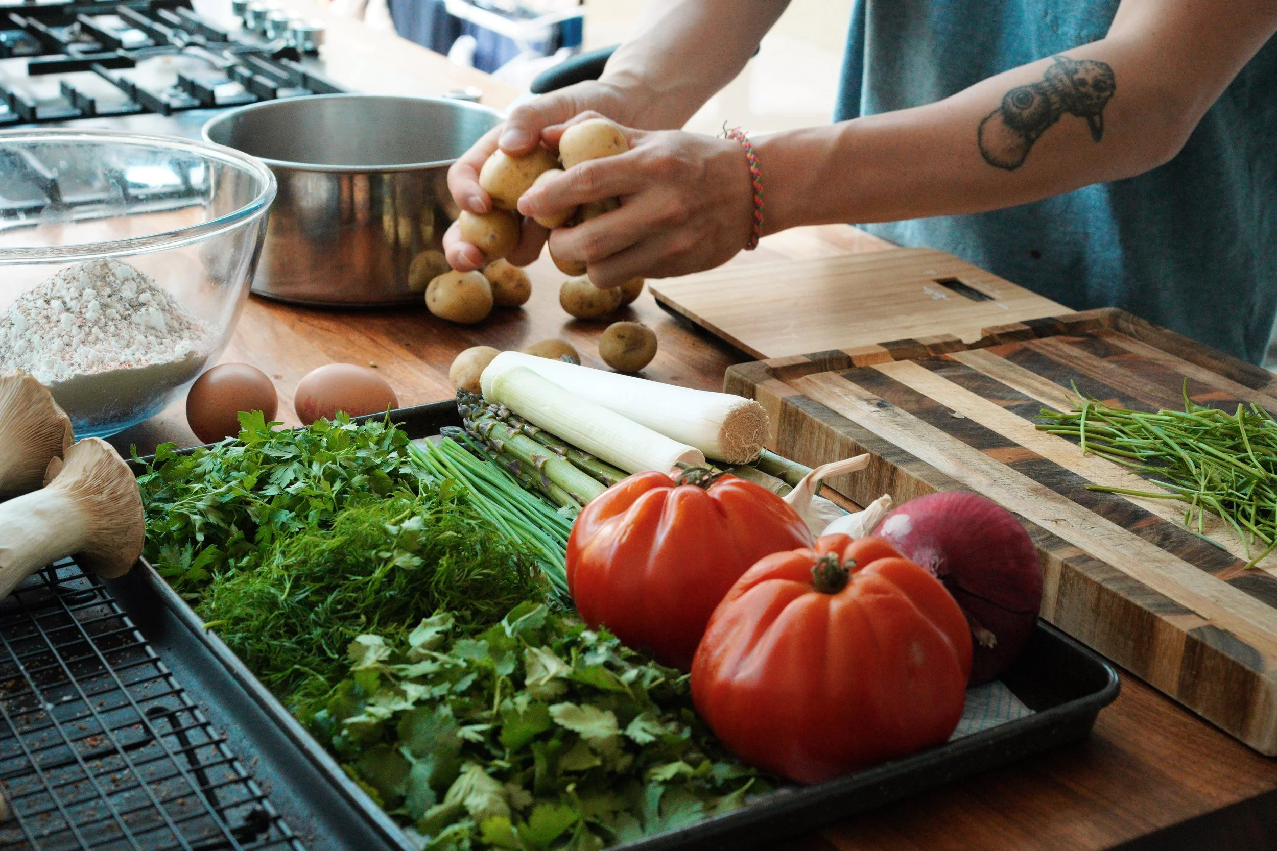 Person preparing vegetables in a kitchen with tomatoes, potatoes, leeks, herbs, and a mixing bowl.