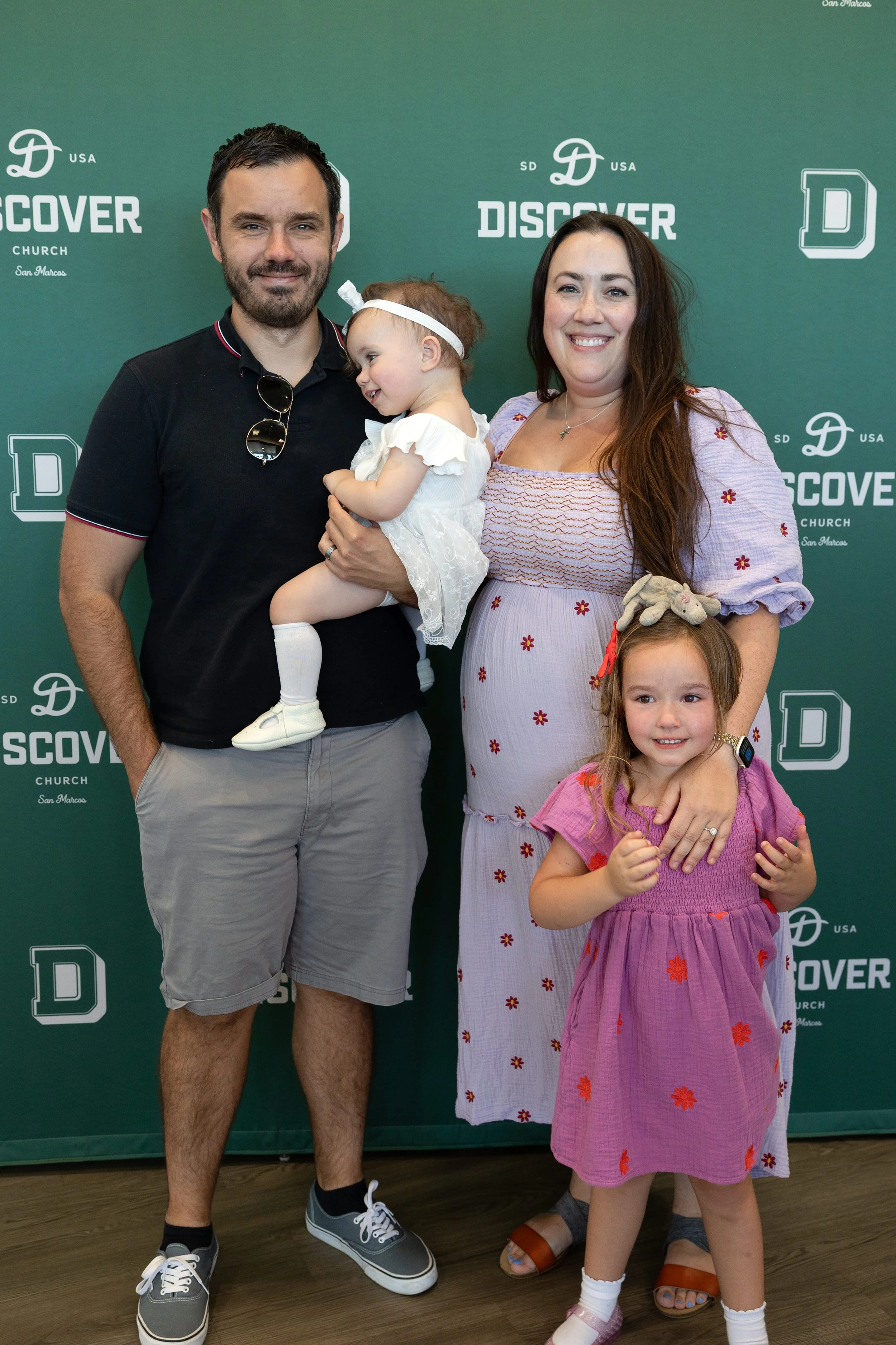 A family of four posing together in front of a green backdrop with 'Discover Church' logos. The father has dark hair and a beard, wearing a black polo shirt, gray shorts, and sneakers. The mother has long brown hair, wearing a pink dress with red floral embroidery. The little girl standing in front has brown hair with a stuffed animal on her head, wearing a pink dress with red floral patterns. The toddler is being held by the father, has light brown hair, and is wearing a white dress and headband.