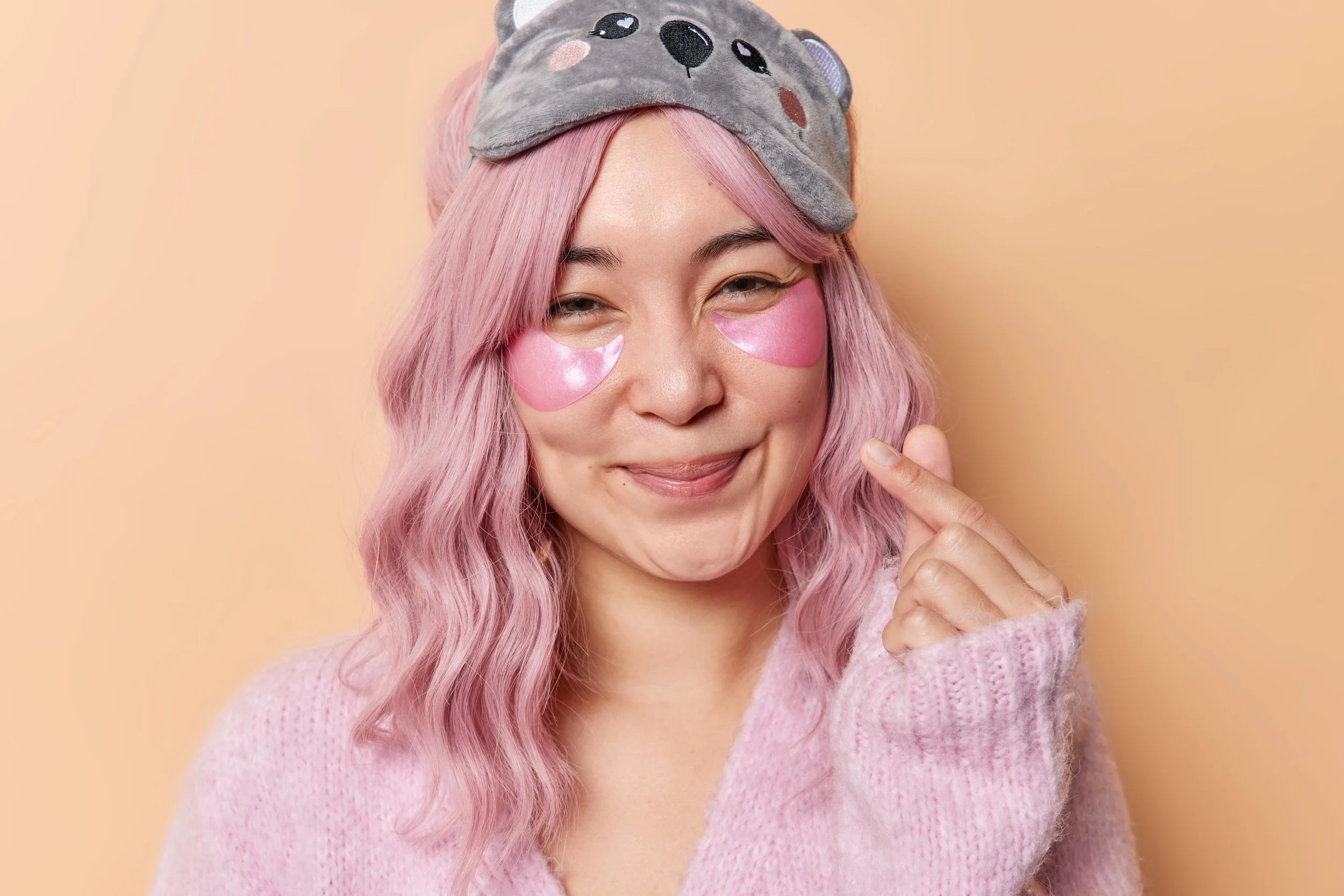 Smiling woman with pink hair wearing hydrogel eye patches and a sleep mask, against a peach background — cozy skincare moment