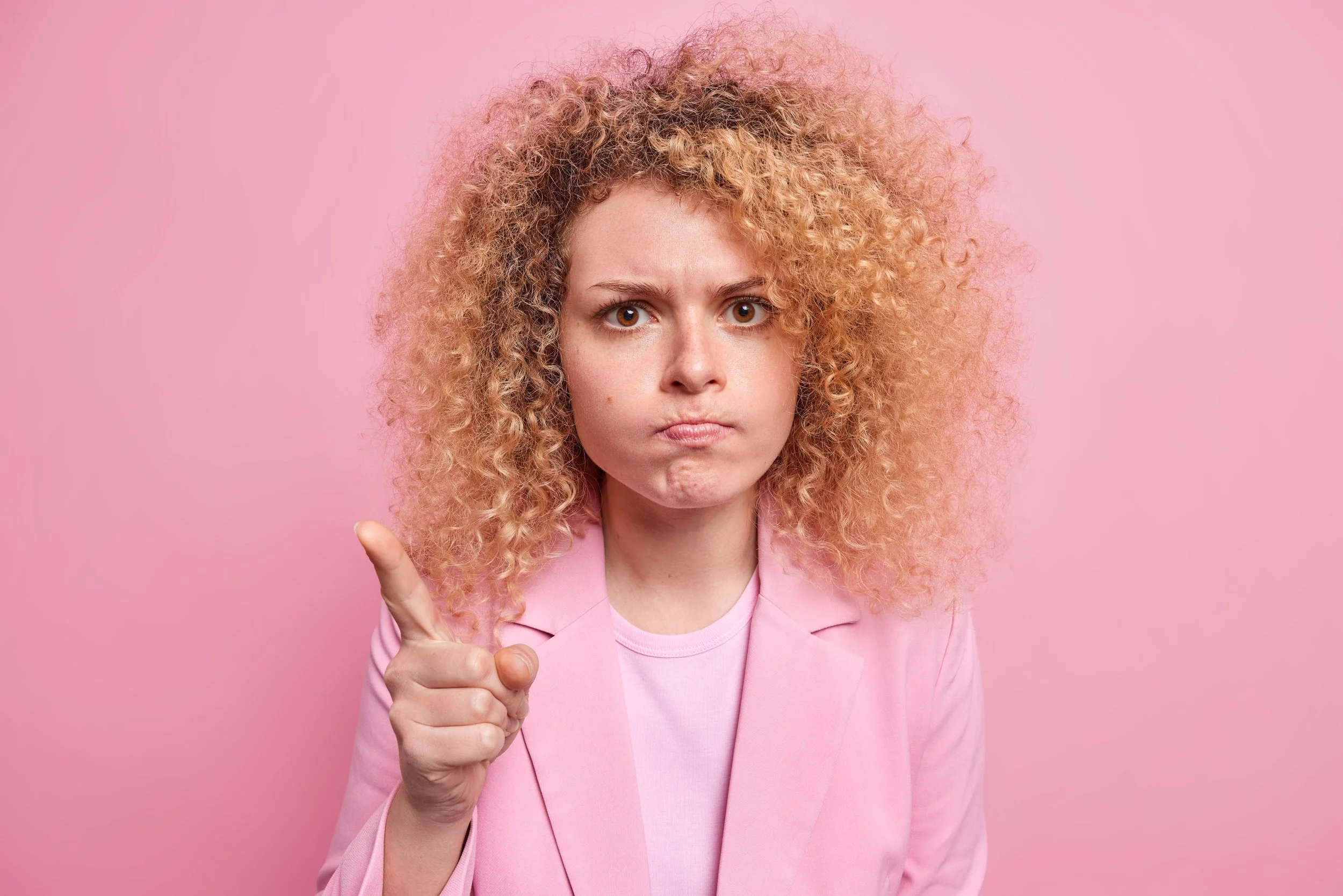 Curly-haired woman pointing and frowning against a pink background, used as a skincare PSA or warning about common barrier-damaging habits.