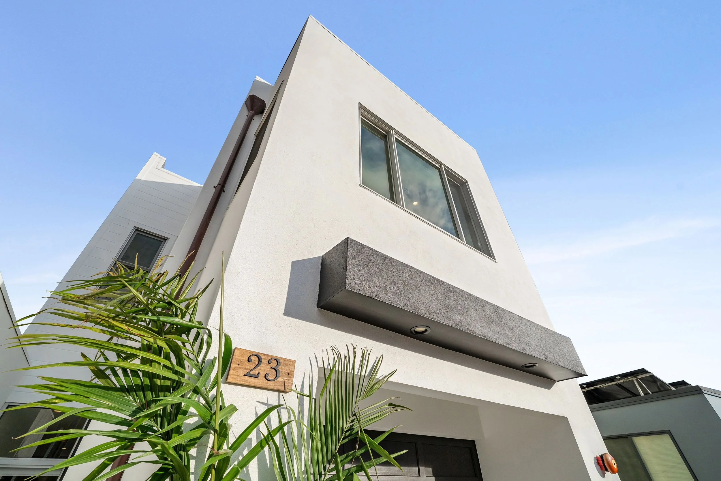Modern white house with large window and house number 23, palm plant in foreground, and blue sky background.