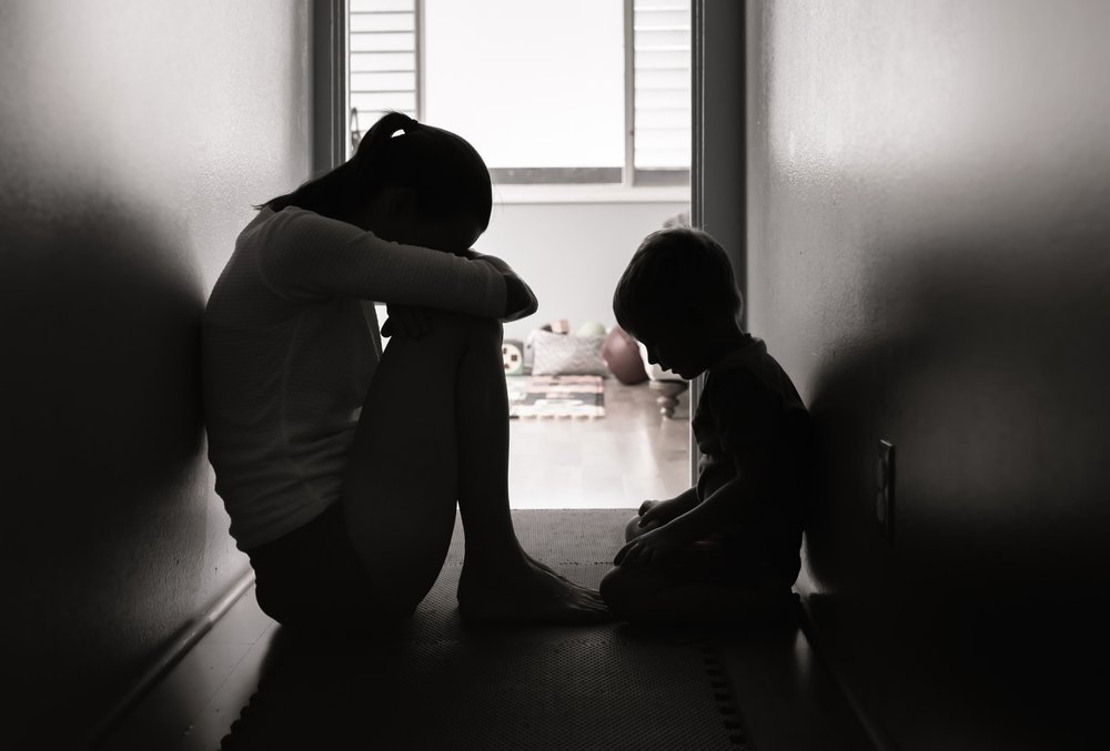 Silhouettes of a woman and a young boy sitting on a hallway floor, facing each other, with a bright window in the background.