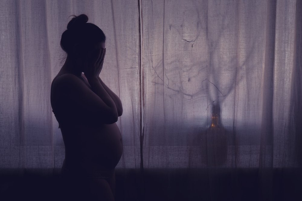 Silhouette of a pregnant woman covering her face, standing next to a window with sheer curtains, with a blurred tree outside.