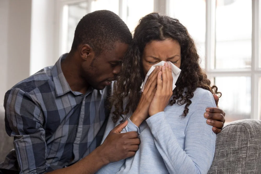 A man comforts a woman who is crying and blowing her nose with a tissue, sitting on a couch in a bright room.