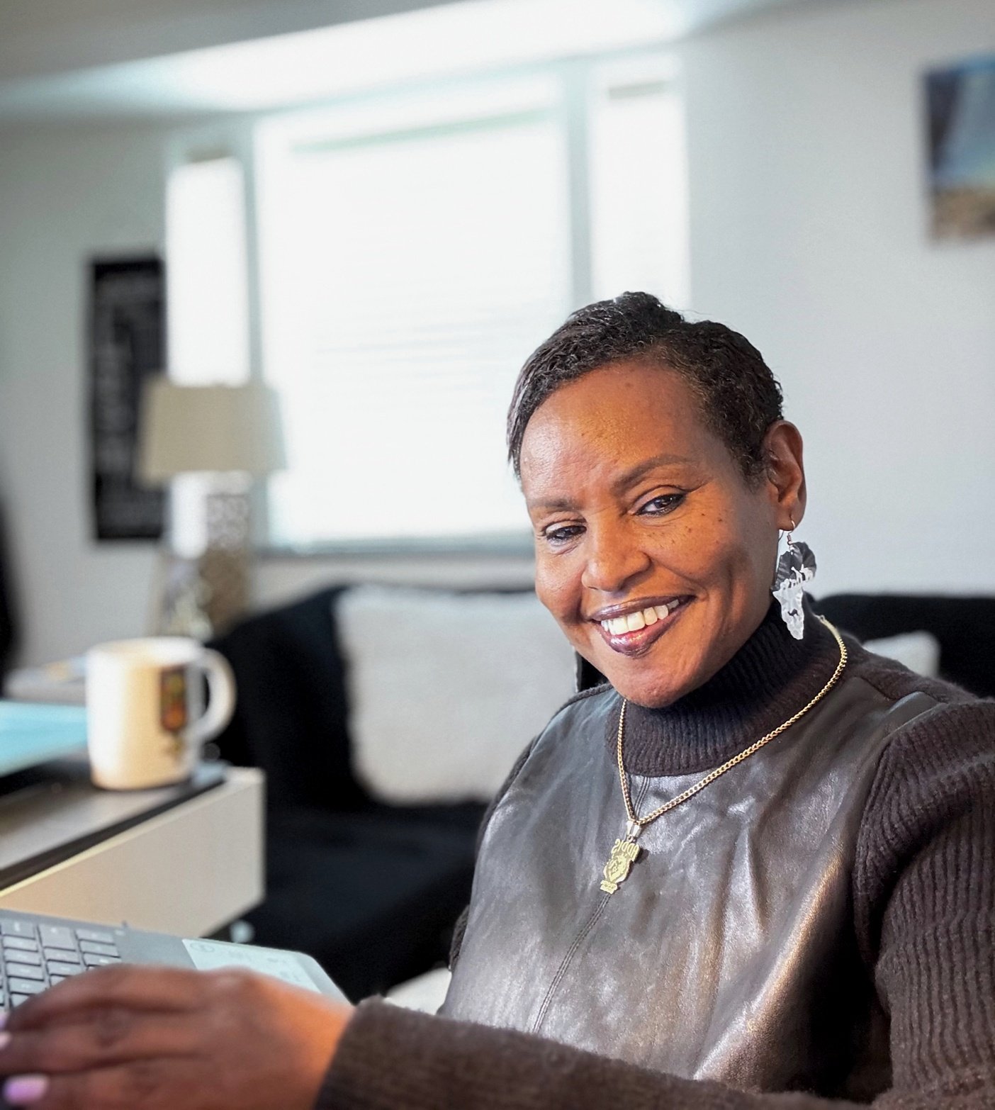 A woman smiling while looking at a phone or tablet in a cozy, well-lit living room with a lamp, mug, and couch in the background.