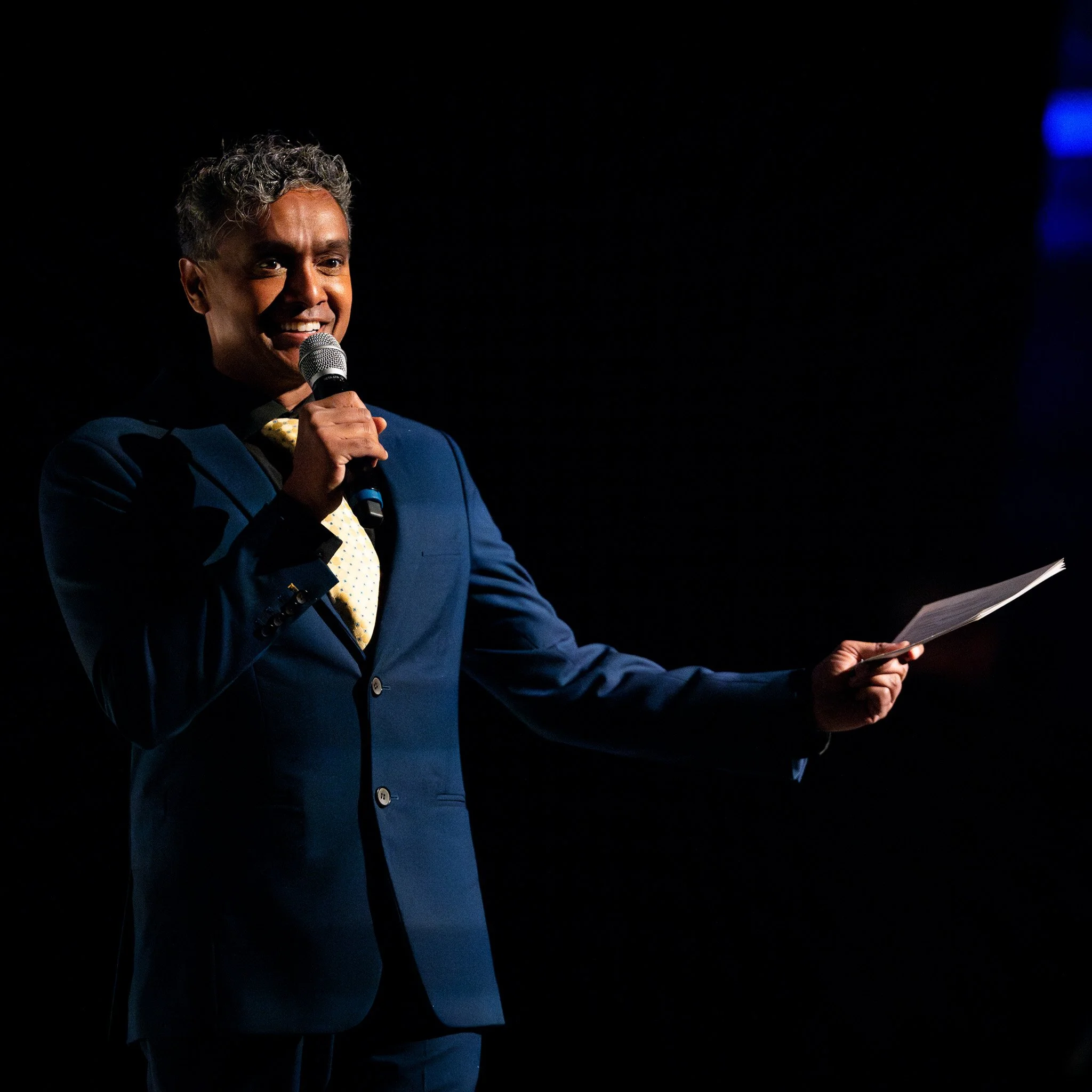 Person in a suit holding a microphone and a piece of paper, speaking on stage.