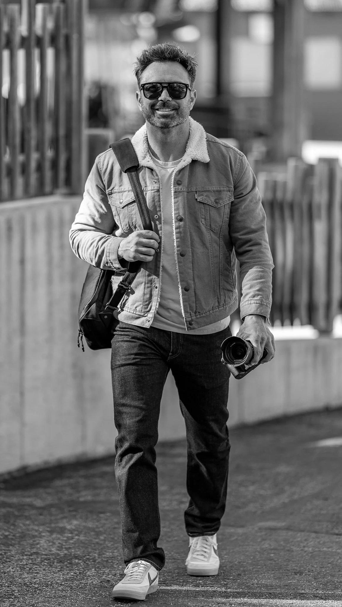 Black and white photo of a man wearing sunglasses, a denim jacket, and jeans, holding a camera in one hand and a bag over his shoulder.