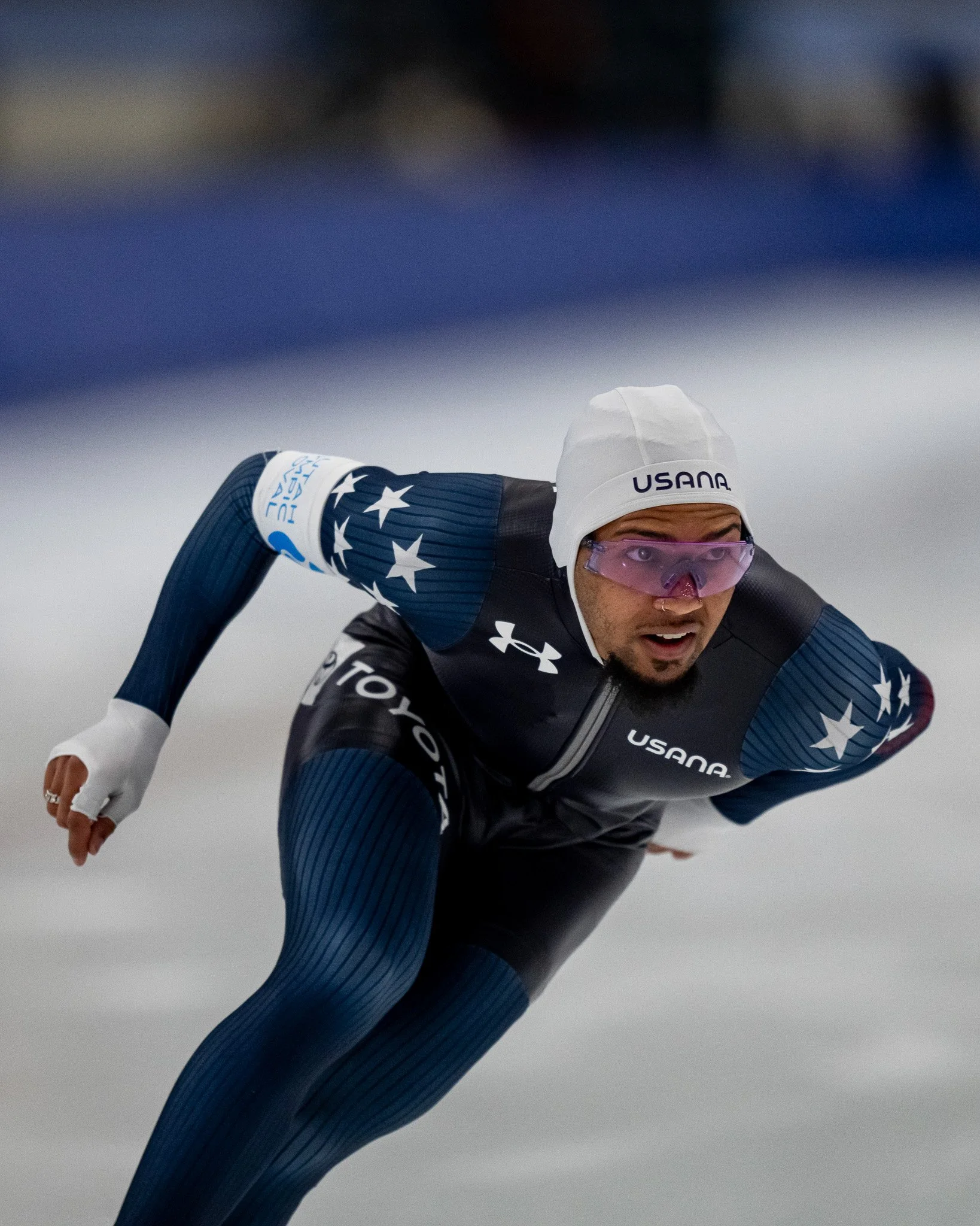 Speed skater in action wearing a dark suit with stars, branded logos, and a white cap.