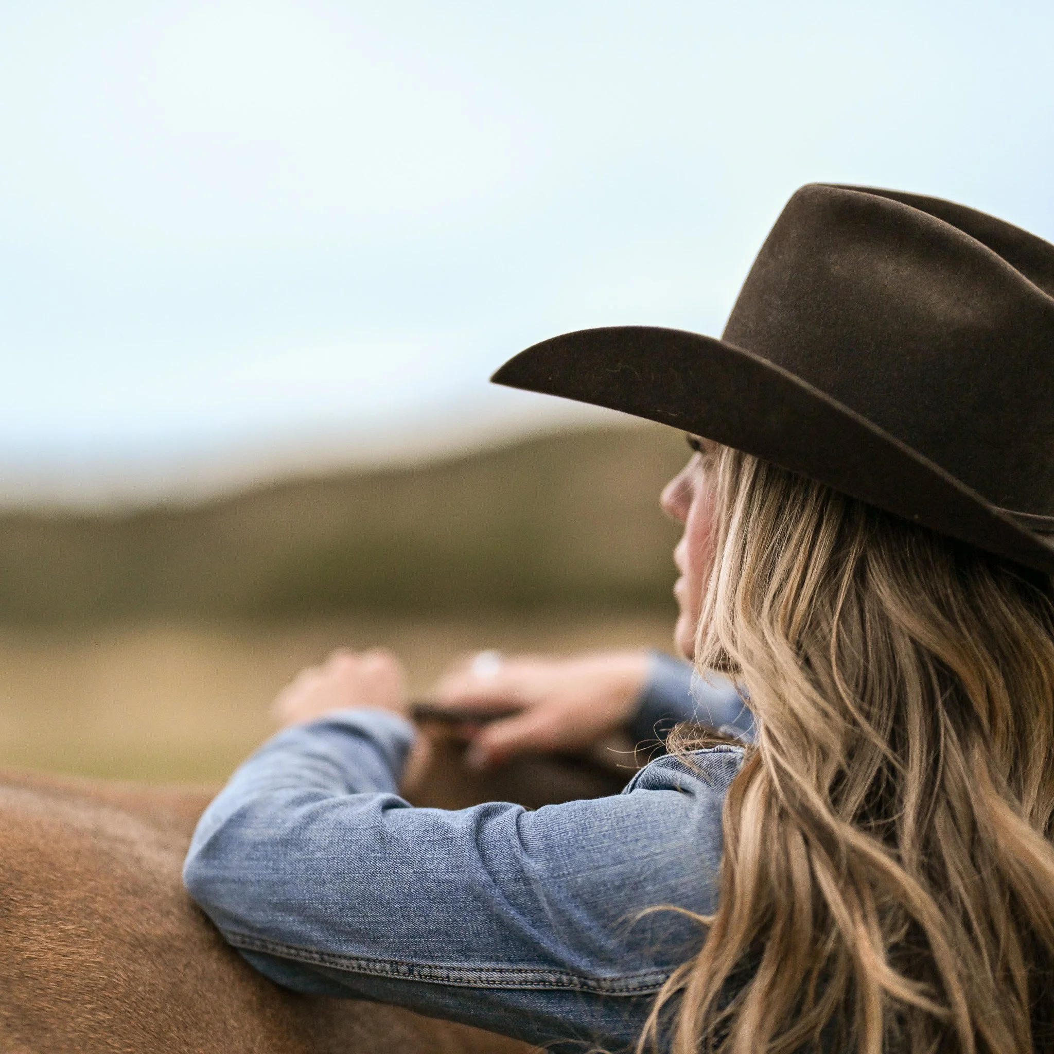 Woman in a cowboy hat and denim jacket sitting outdoors, looking at the horizon.