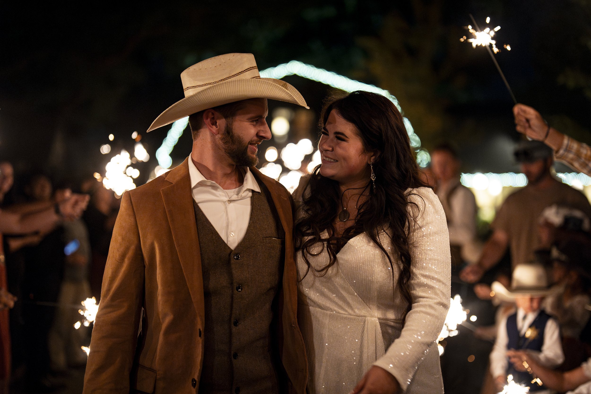 A couple at an outdoor nighttime celebration, with the man wearing a cowboy hat and brown suit, and the woman in a white dress. They are surrounded by people holding sparklers.
