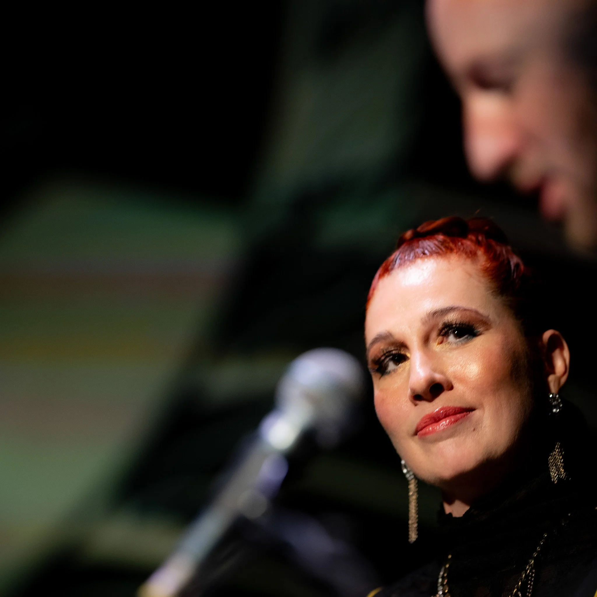 Woman with red hair near microphone on stage, blurred background.