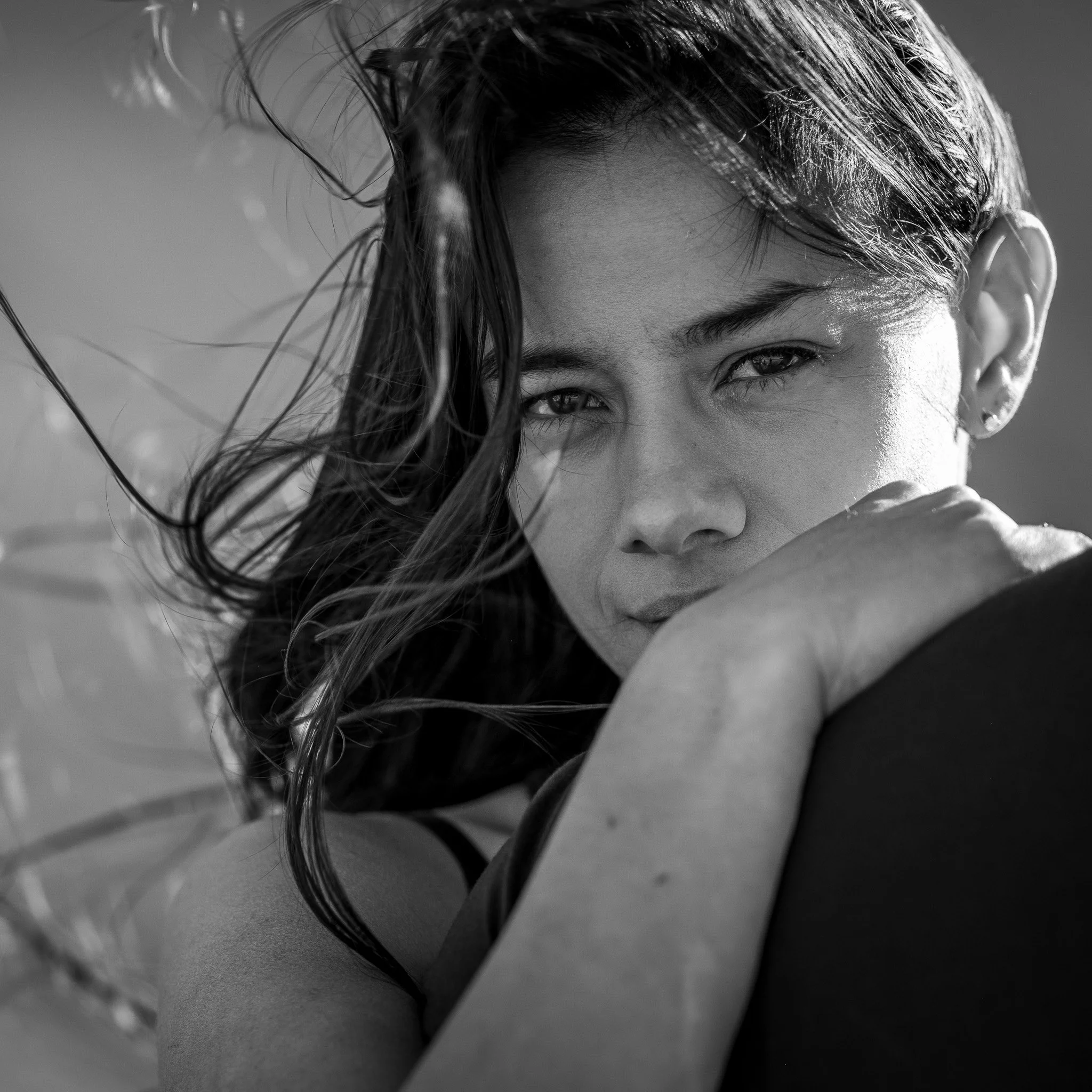 Black and white portrait of a woman with windblown hair, resting her chin on her hand, looking directly at the camera.
