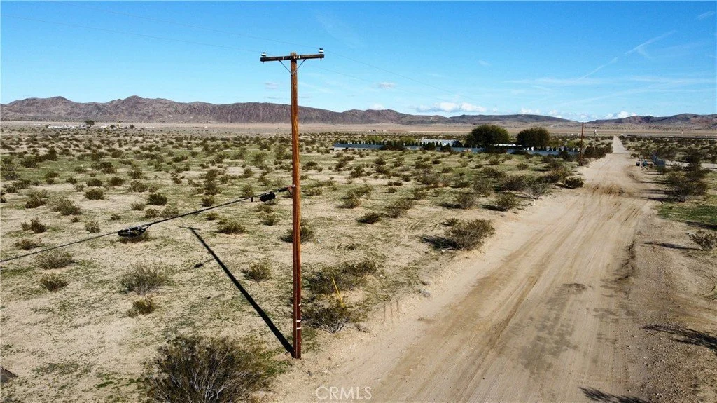 A rural dirt road with a utility pole on the left side and sparse desert vegetation. In the distance, there are mountains, trees near a body of water, and a line of cars along the road.